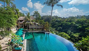 A infinity pool with a clear blue water edge, surrounded by lush greenery and a thatched-roof building. - Pita Maha (Ubud)