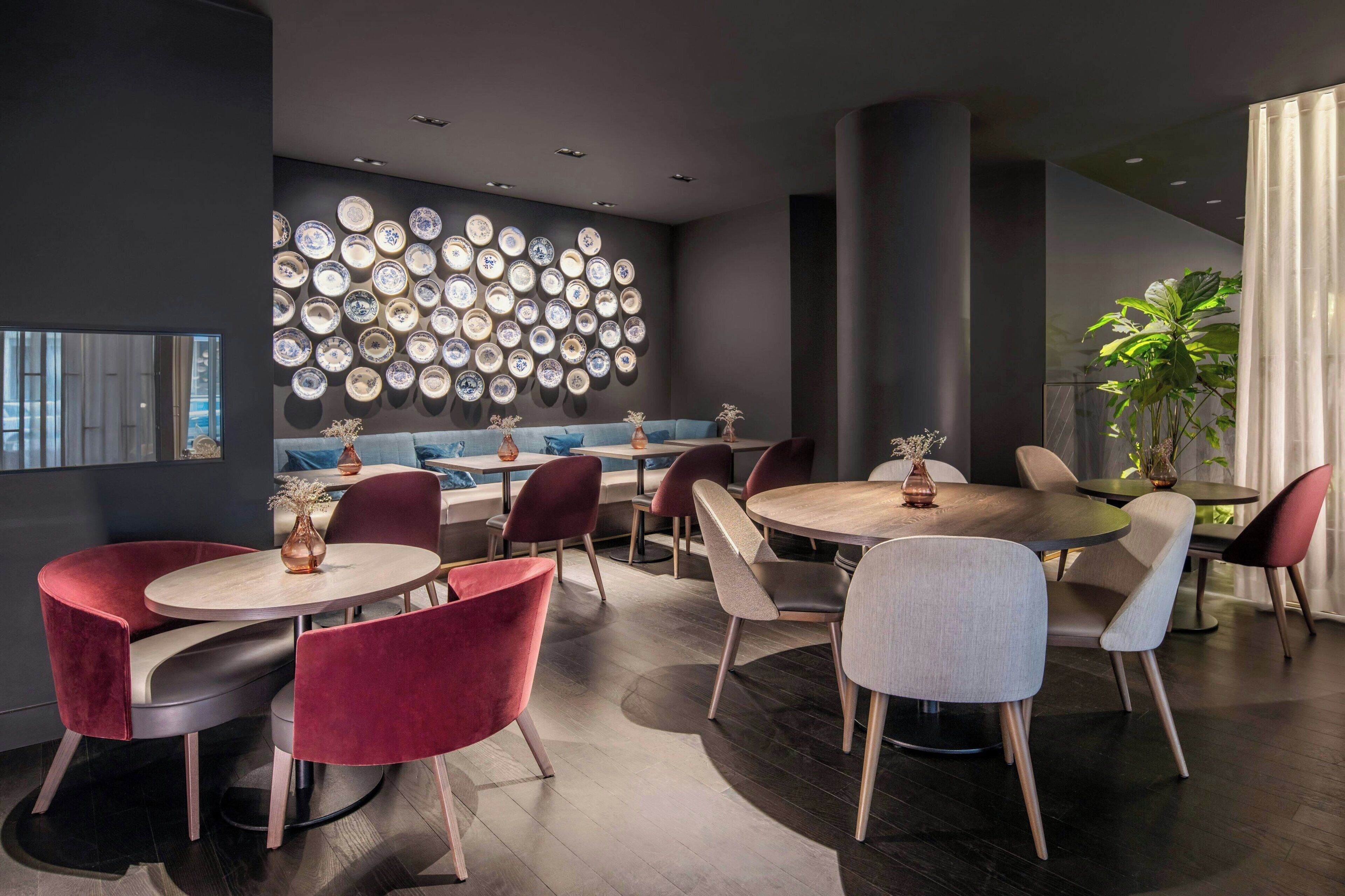 Un comedor moderno con mesas y sillas redondas, una pared decorada con platos y una planta grande en maceta.