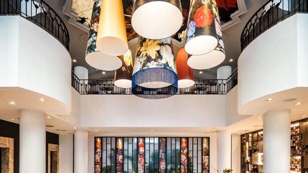 A spacious lobby with a high ceiling, large hanging lamps, a seating area with sofas and chairs, and a bookshelf filled with books.
