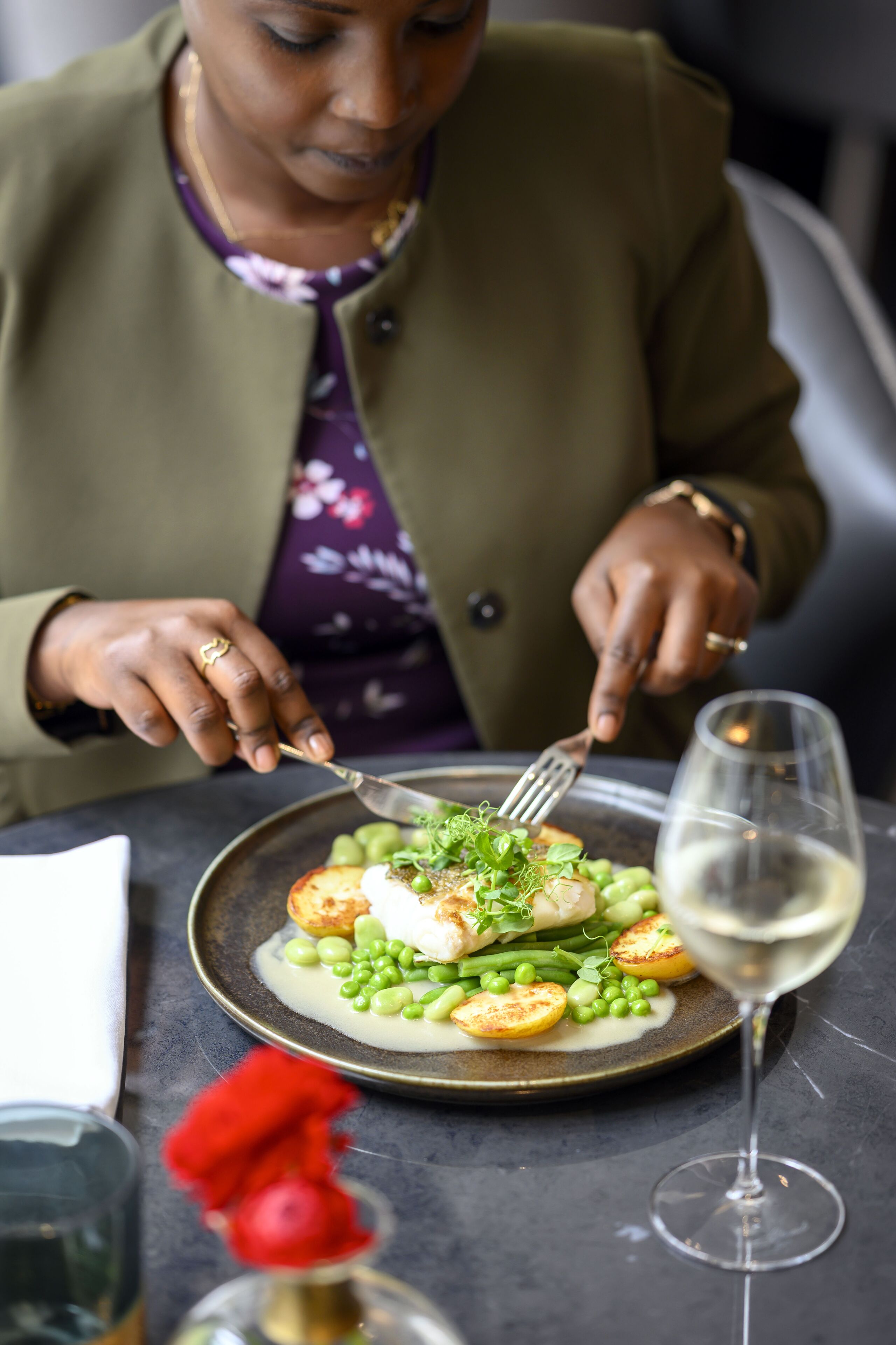 Iemand eet aan een tafel met een bord eten, een glas witte wijn en een vaas met een rode bloem.