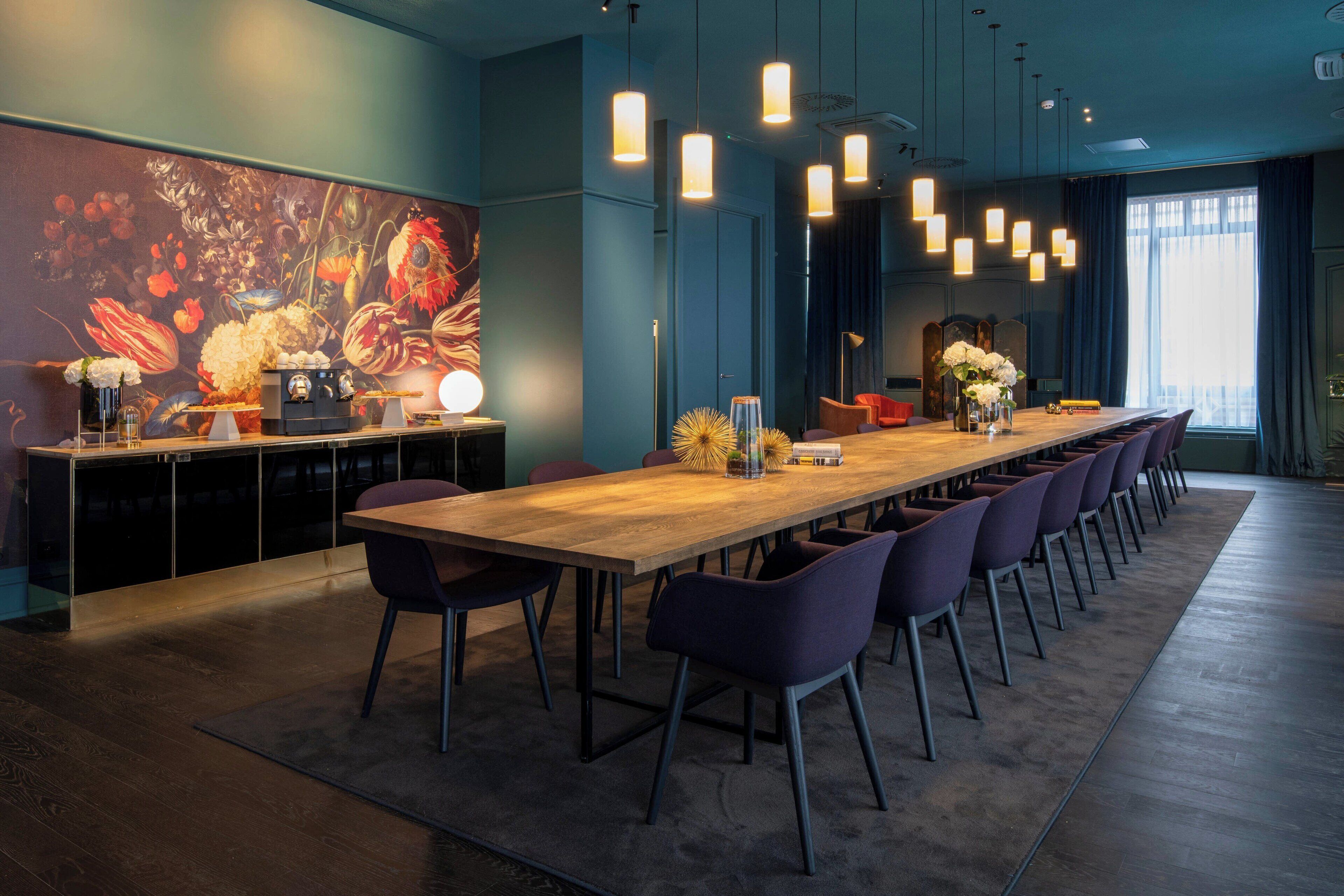 A modern conference room with a long table, purple chairs, pendant lighting, and a floral wall art piece.