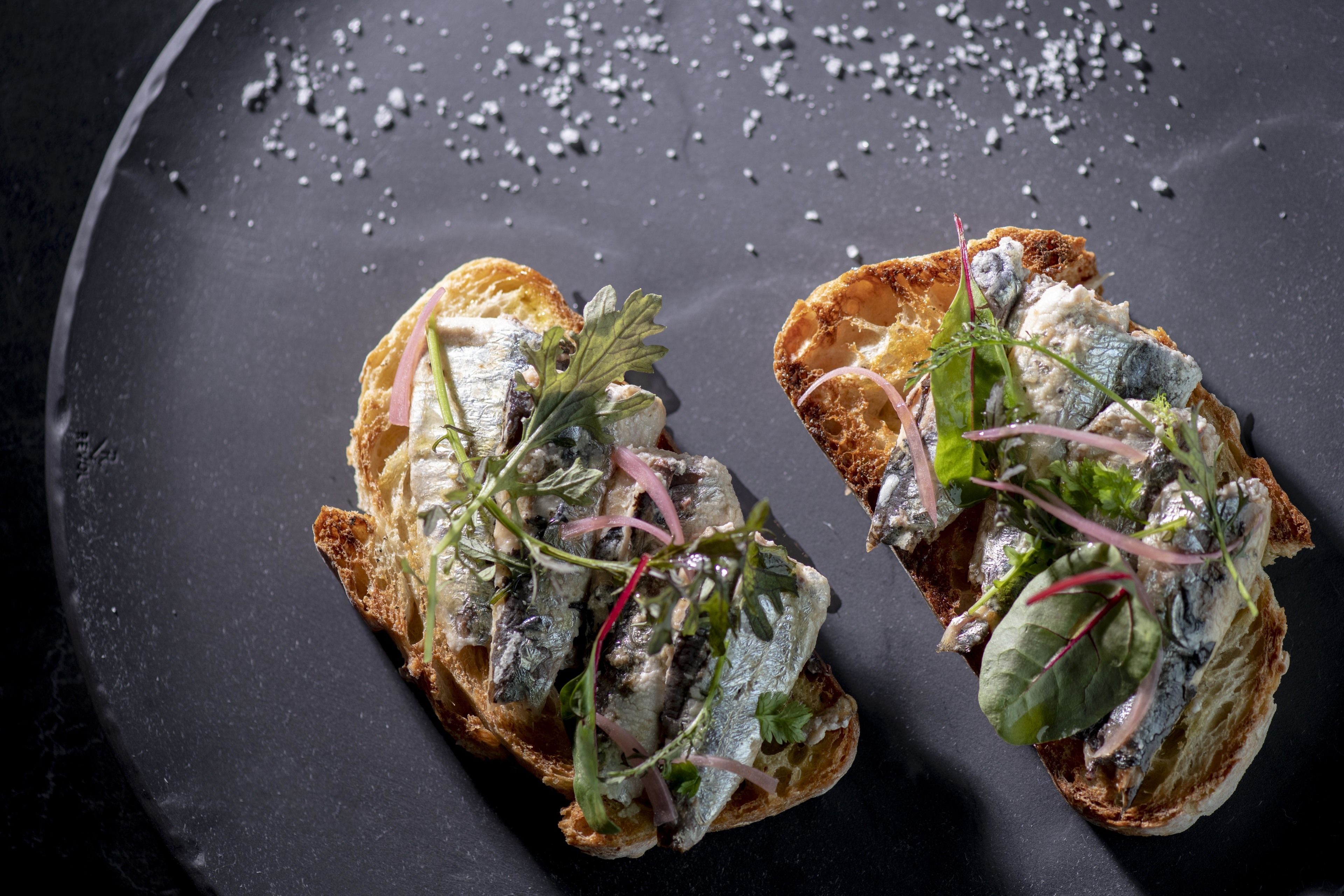 Two pieces of toast topped with fish, greens, and onions on a dark plate.
