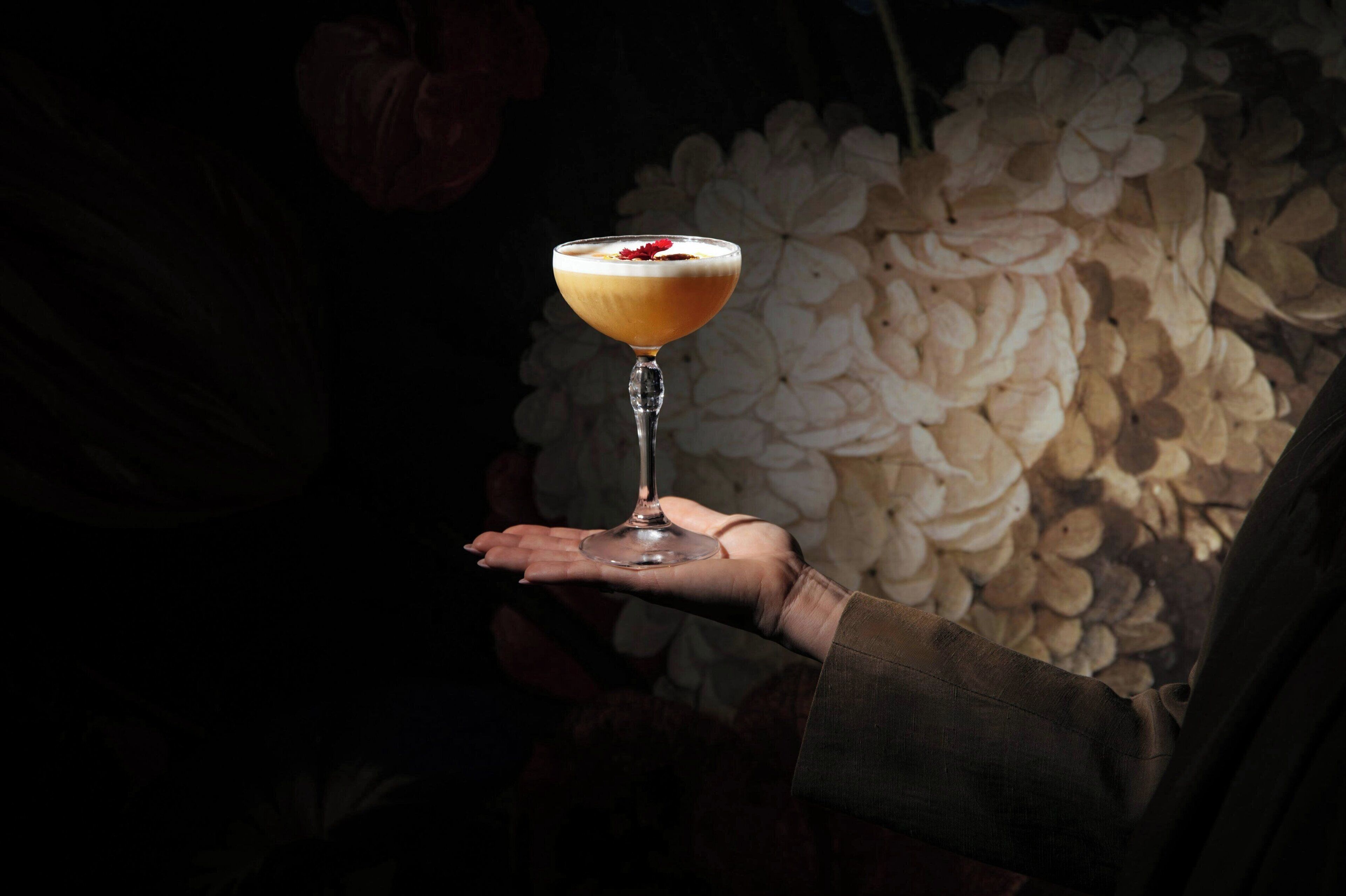 A hand holding a cocktail glass with a creamy drink and a garnish, set against a backdrop of white flowers.