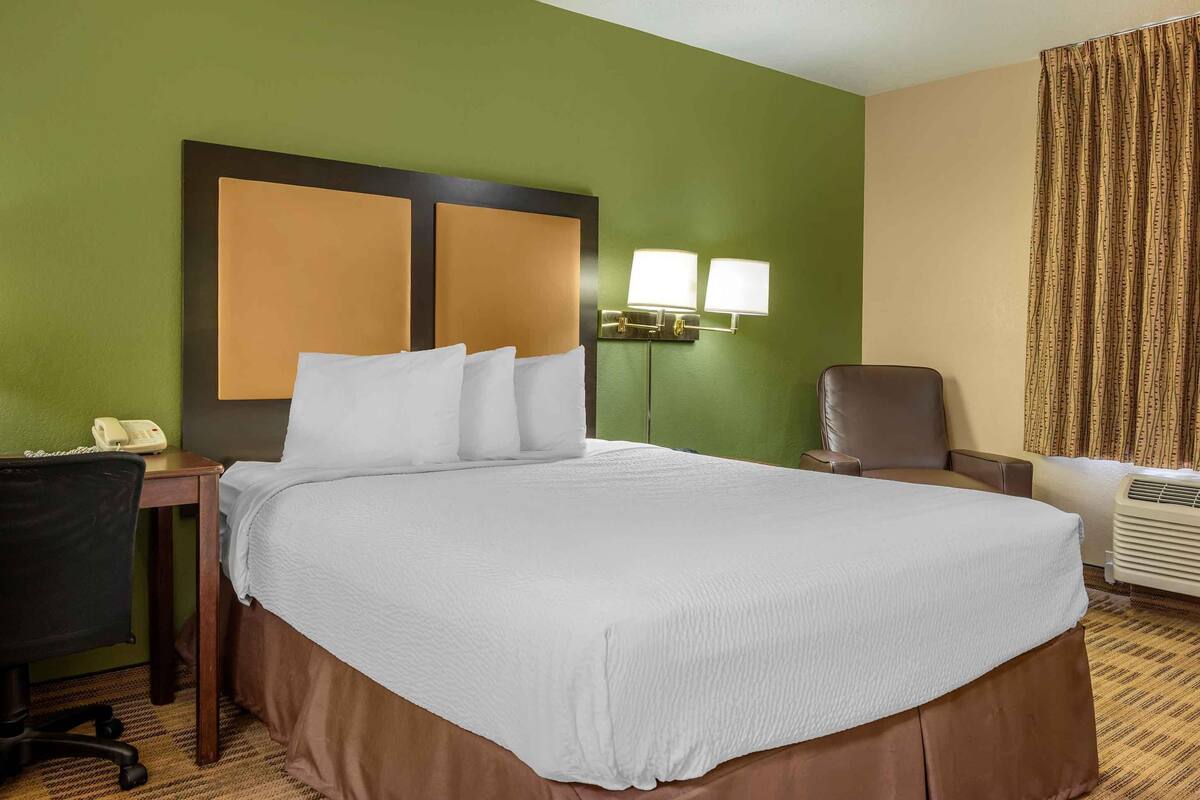 A hotel room with a large bed, a desk with a telephone, a chair, and a wall-mounted lamp.