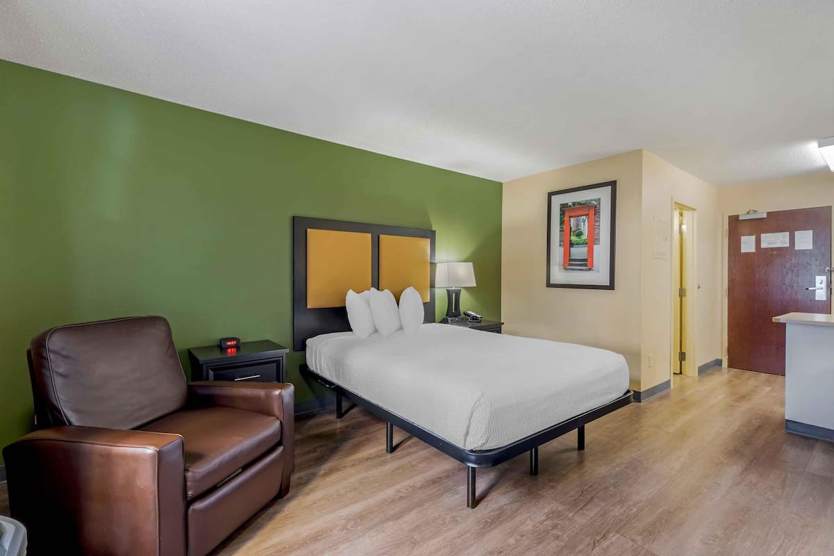 A hotel room with a bed, a chair, a nightstand, a lamp, and a door.