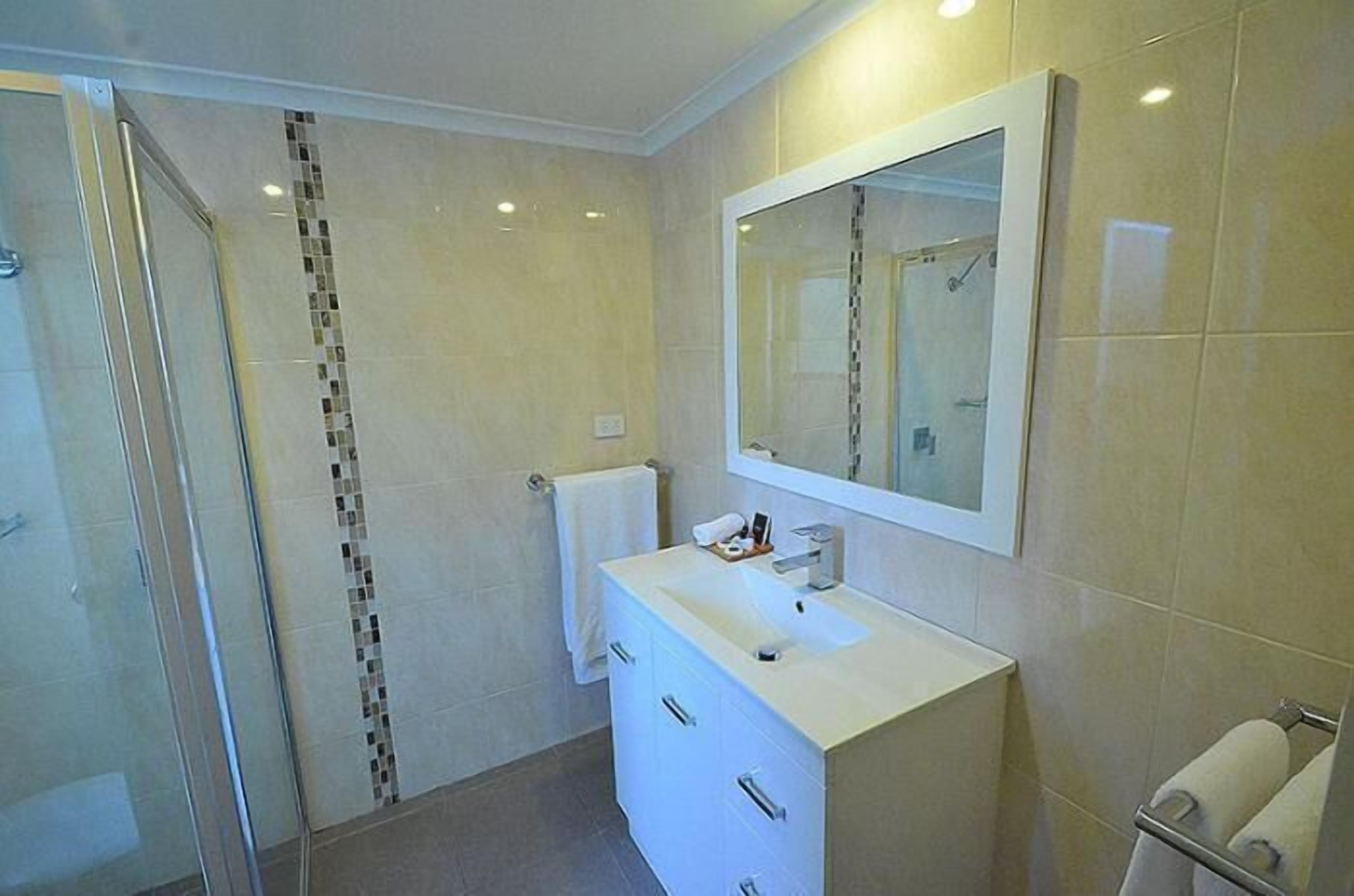 Un baño moderno con cabina de ducha de vidrio, tocador blanco con espejo y suelo de baldosas.