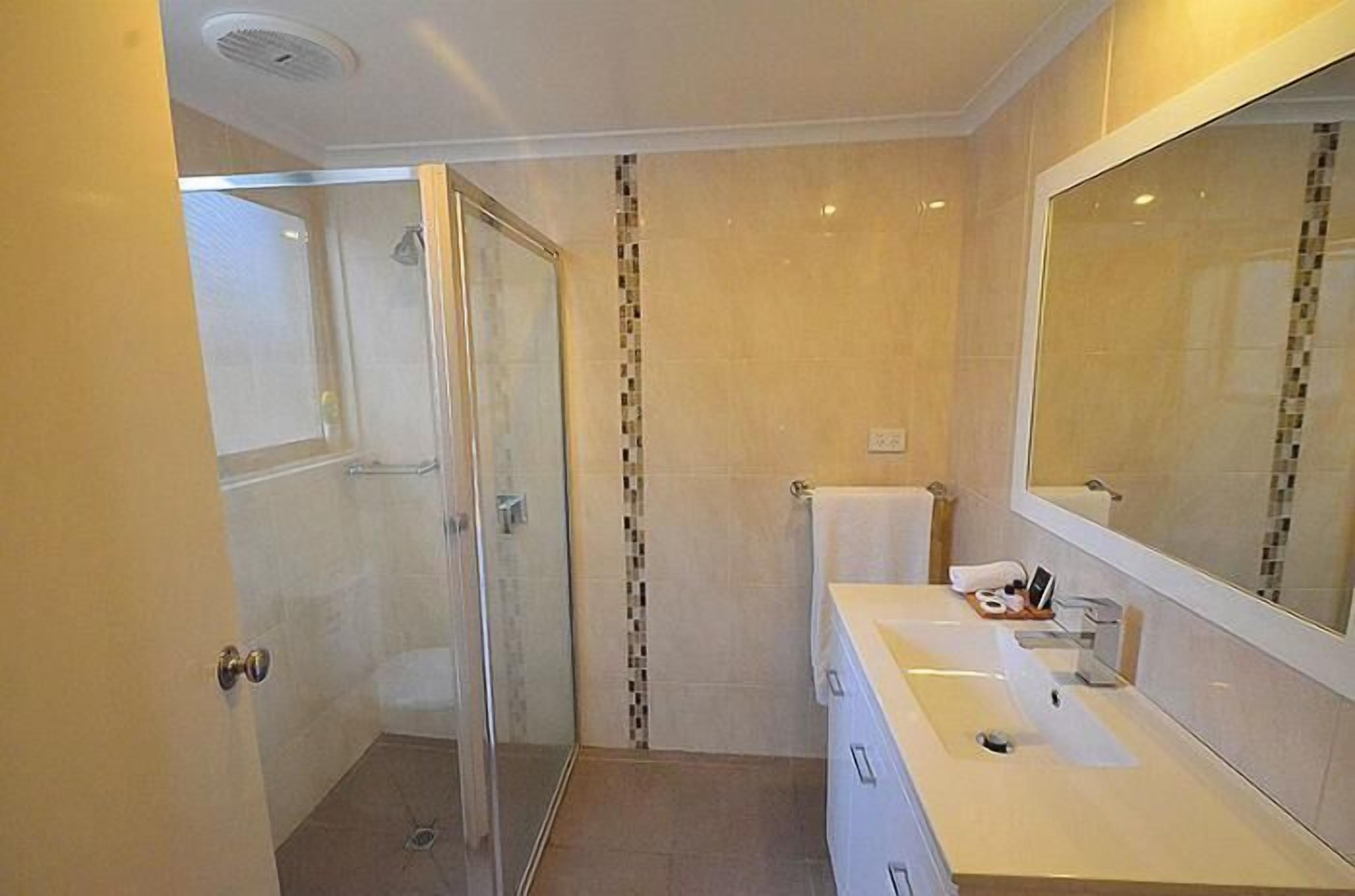 Un baño moderno con cabina de ducha de vidrio, un lavabo blanco con espejo y paredes con azulejos color beige.