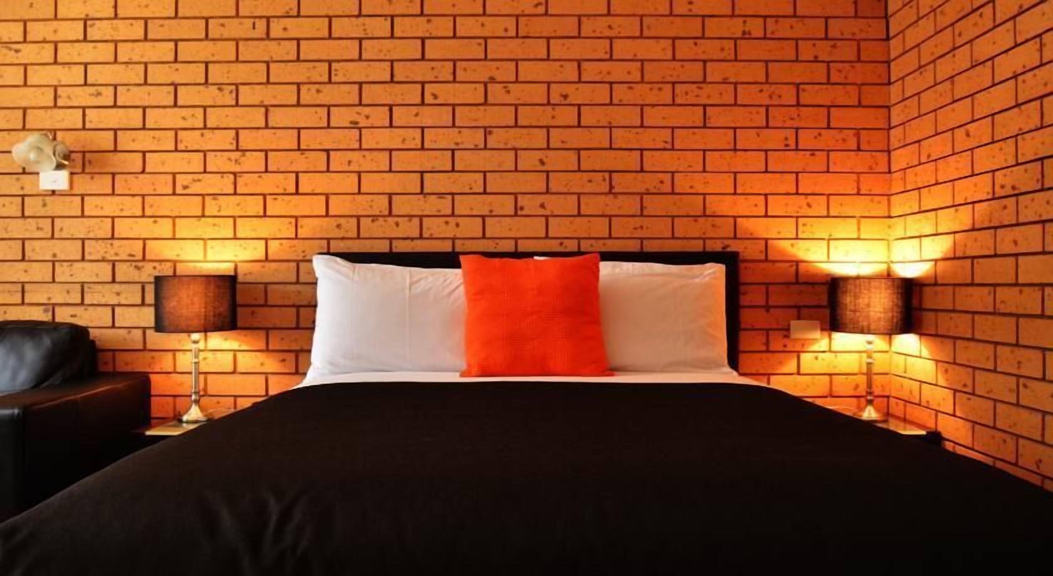 A hotel room with a brick-patterned wall, a black bed with white and orange pillows, and two bedside lamps.