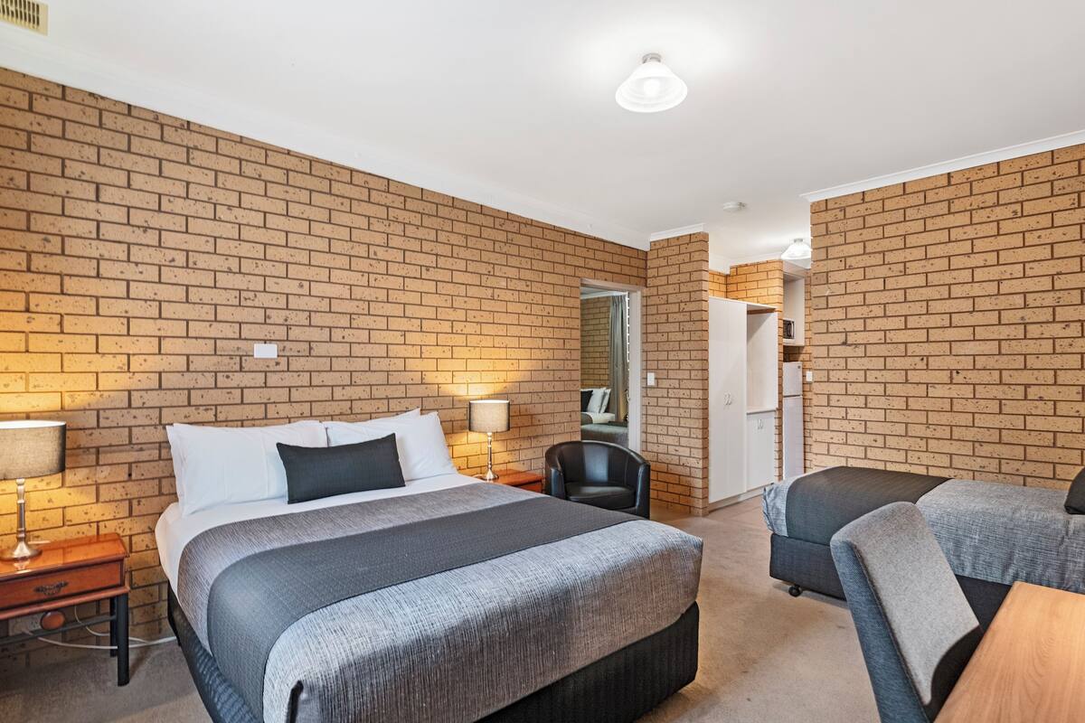 A hotel room with a brick wall, a large bed with a grey blanket, a desk with a lamp, and a chair.