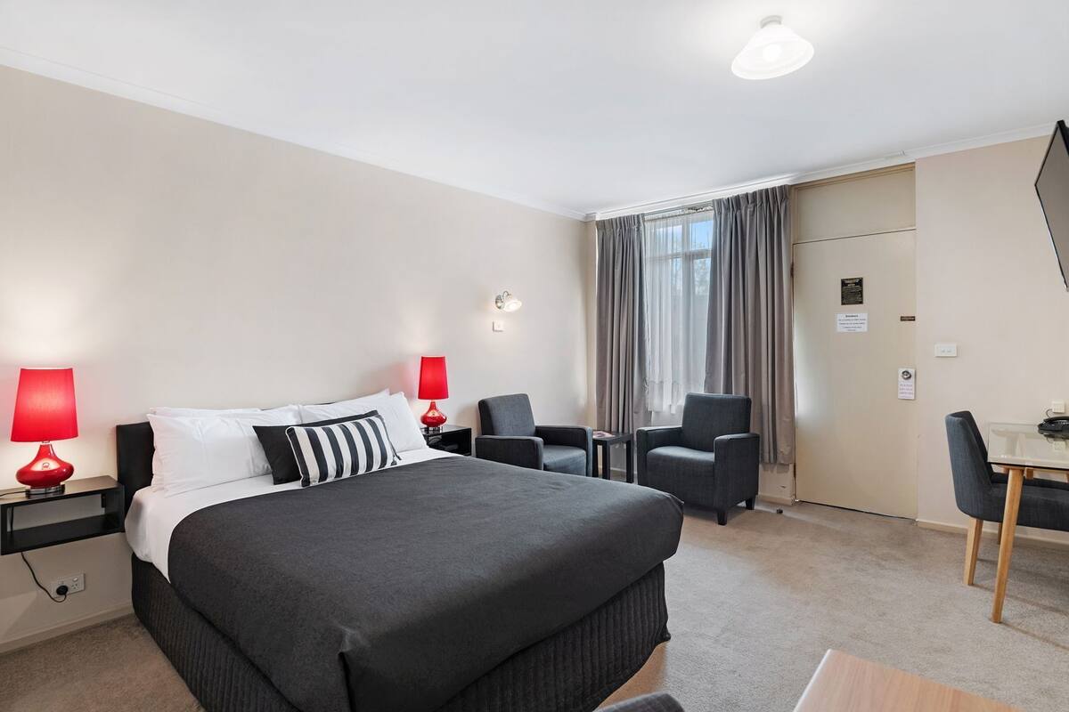 A hotel room with a large bed, two bedside tables with lamps, a desk with a chair, and a window with curtains.