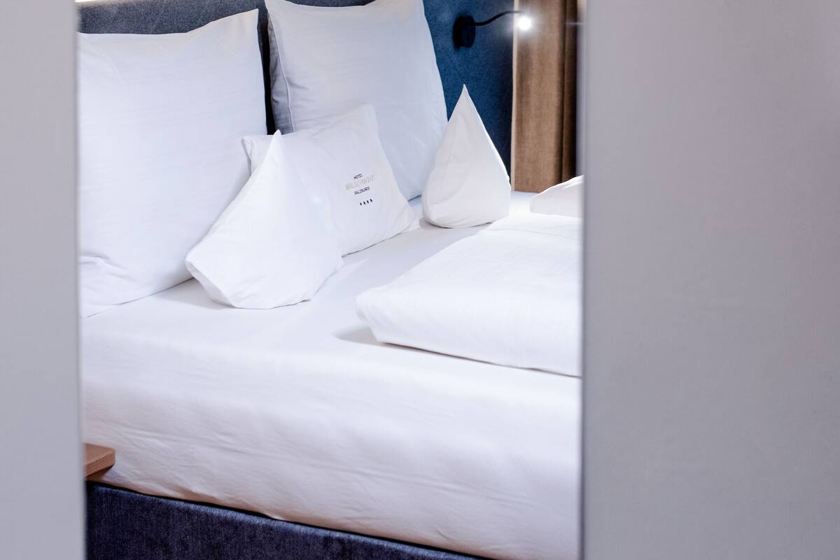 Una cama bien hecha con sábanas y almohadas blancas, un cabecero oscuro y una lámpara de pared.