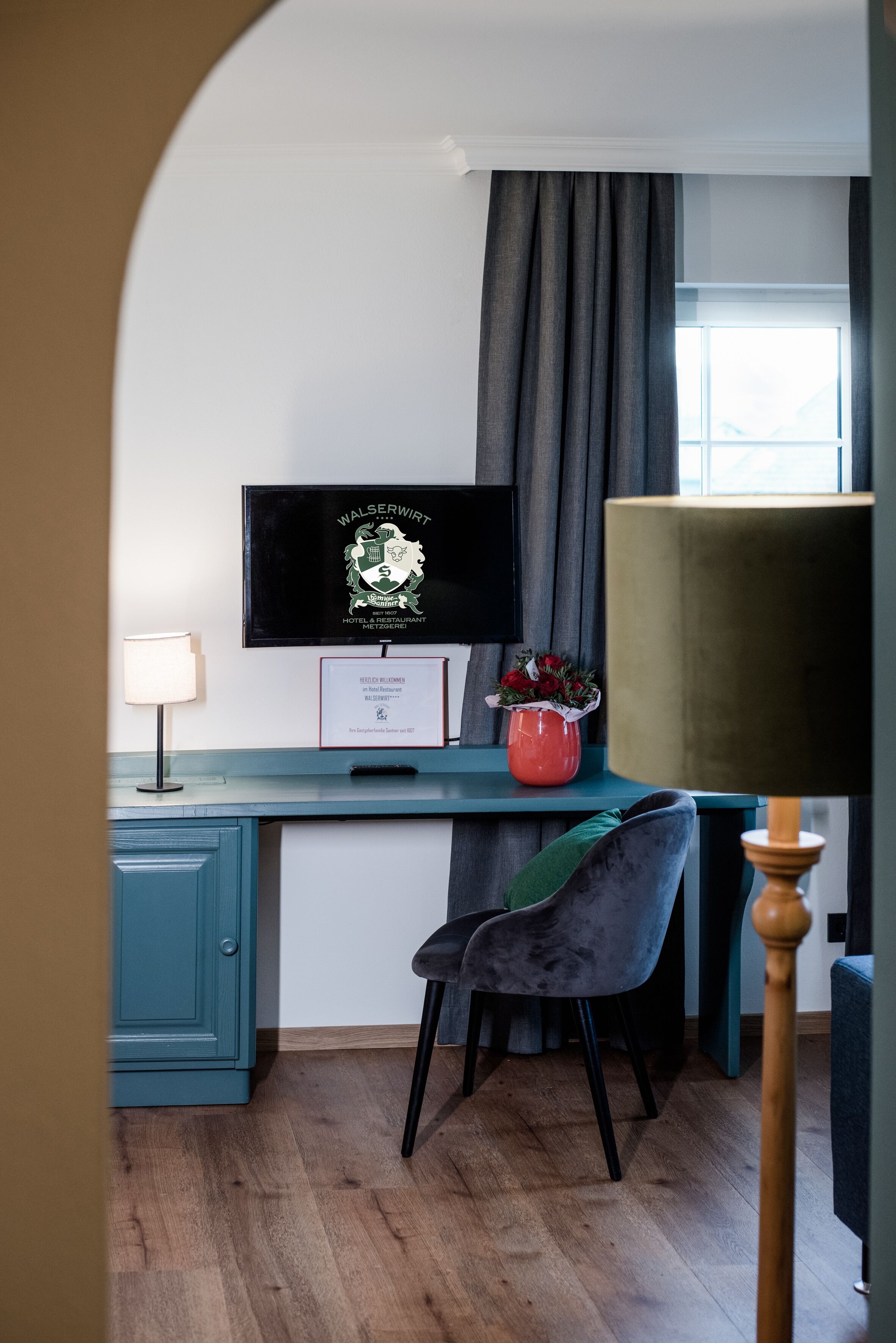A room with a teal desk, a television mounted on the wall, a red vase with flowers, a grey chair, and a lamp.