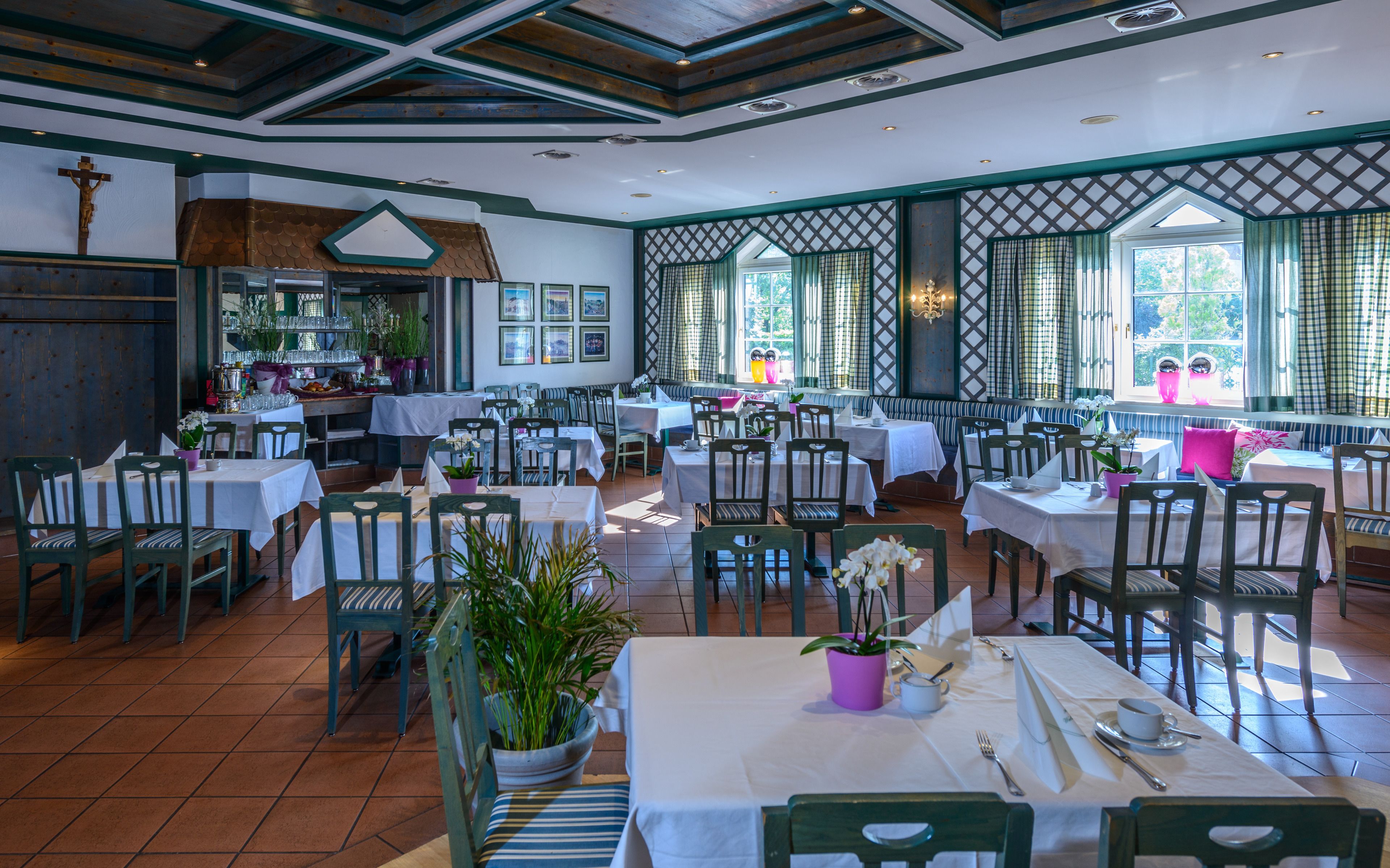 A restaurant with multiple tables and chairs, a wooden ceiling, and large windows with curtains.