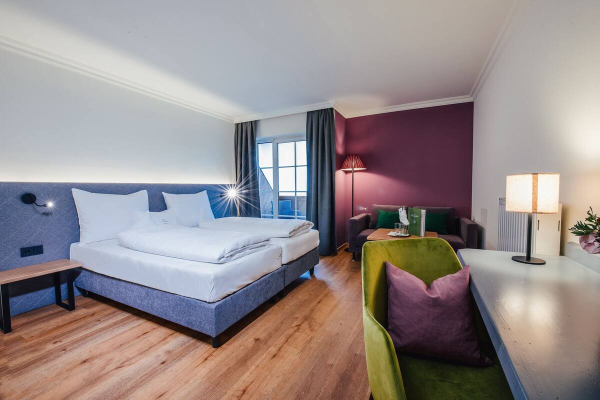 Habitación de hotel con cama, un banco, un sillón verde, un escritorio con lámpara y una ventana con cortinas.