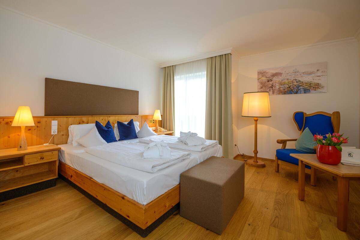 Una habitación de hotel con una cama de madera, dos mesitas de noche con lámparas, una silla, una pequeña mesa con un florero y un cuadro en la pared.