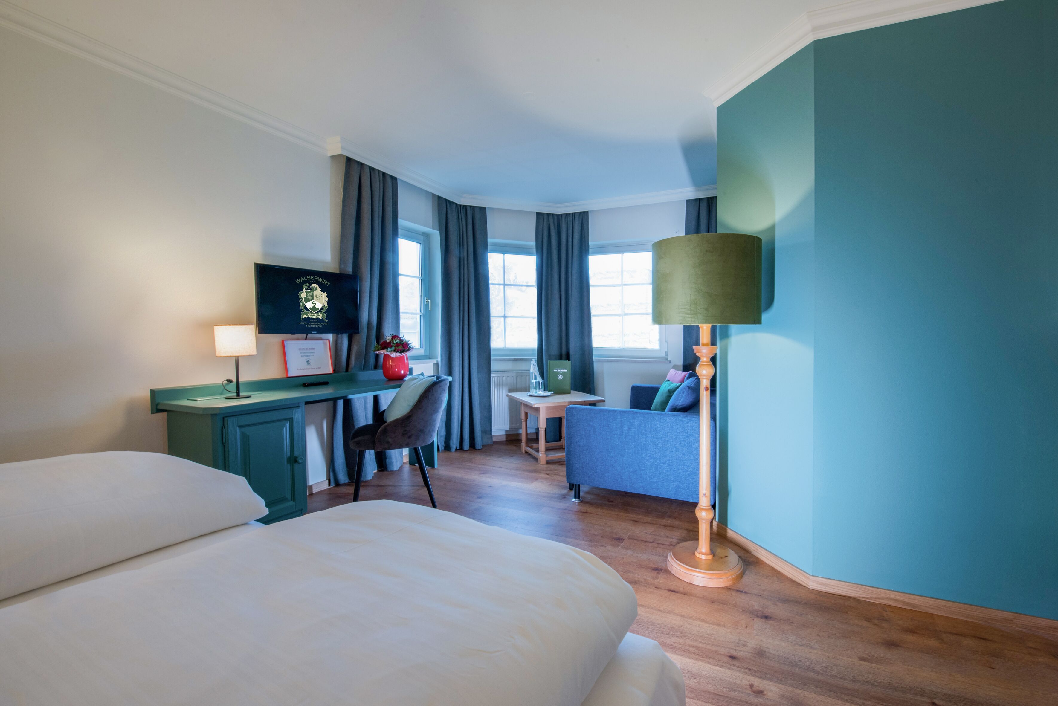 A hotel room with a bed, a desk with a computer, a chair, a small table, and a lamp.