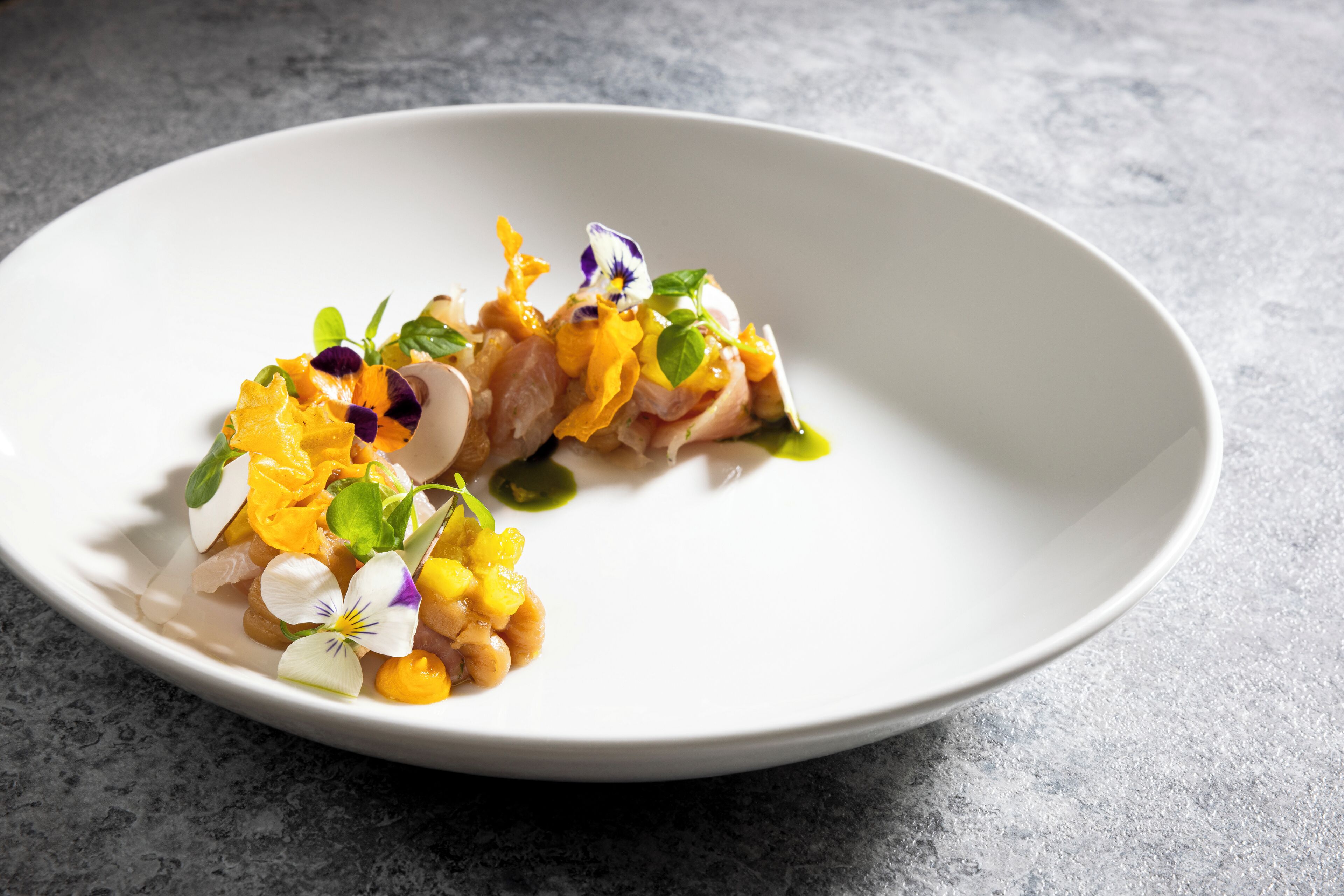Une assiette blanche contenant un plat composé de poisson et de légumes, posée sur une surface grise texturée.