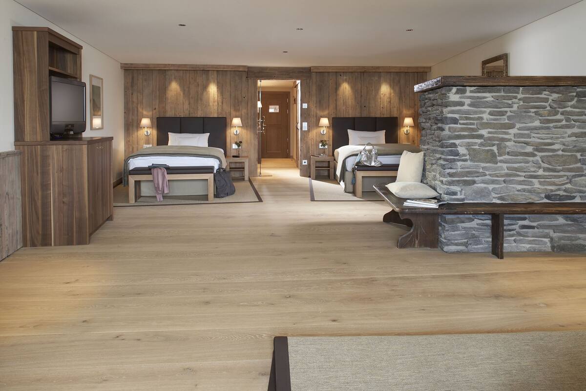 Una habitación de hotel moderna con suelo de madera, dos camas, una chimenea de piedra y un televisor sobre un mueble de madera.