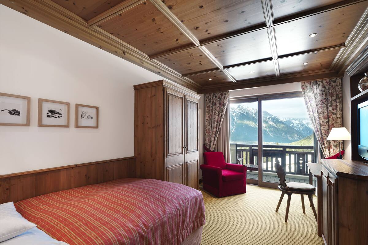Habitación de hotel con una cama, una silla roja, un escritorio y un ventanal con vistas a la montaña.