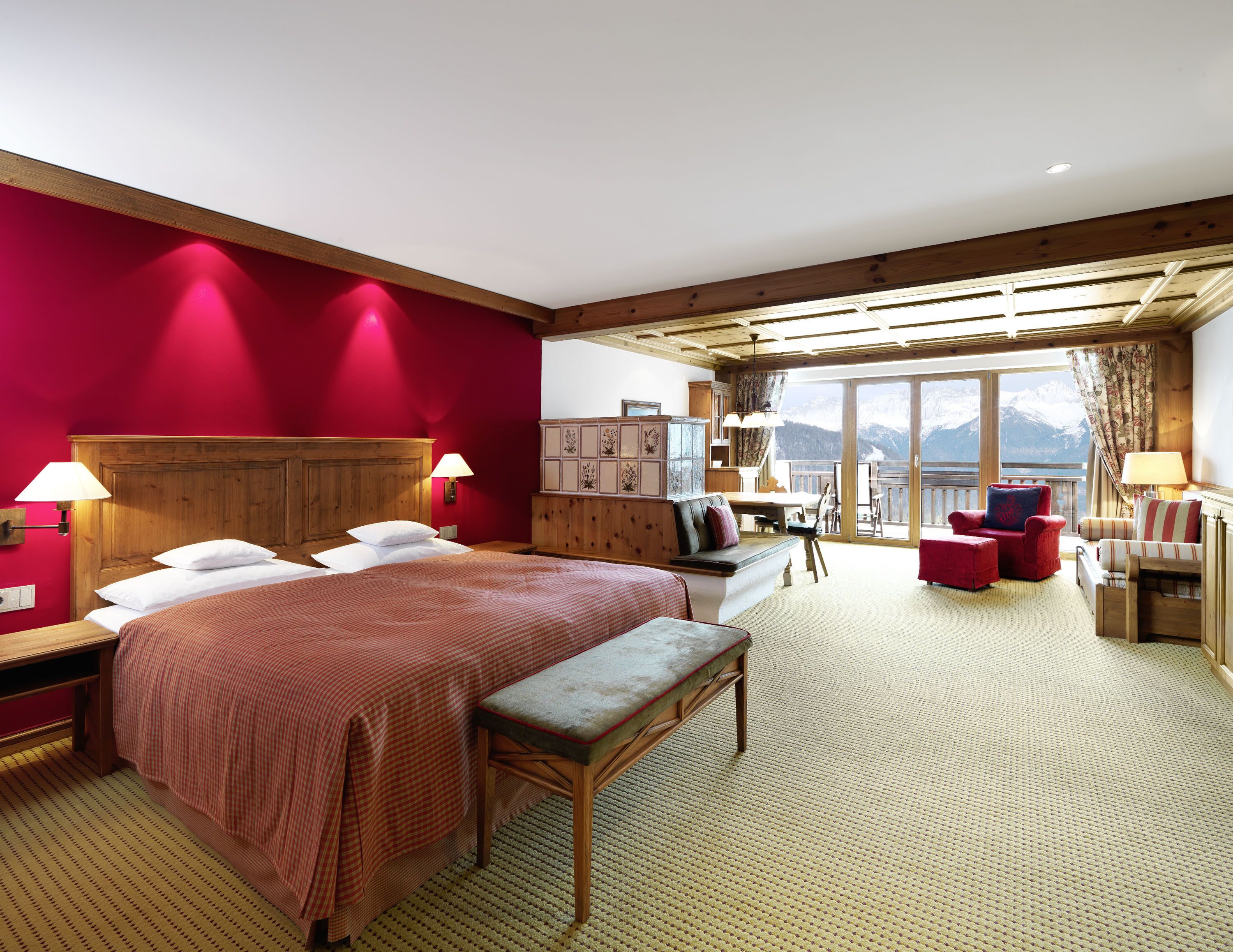 A hotel room with a large bed, a wooden bench, a desk, and a view of mountains through the windows.