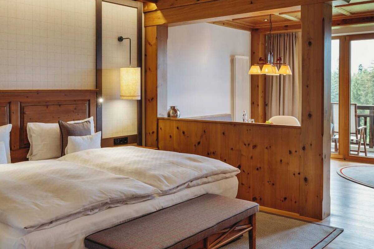 Un dormitorio con cabecera de madera, una cama grande con ropa de cama blanca, un banco al pie de la cama, una mesita de noche con lámpara, y vistas al exterior a través de una puerta corredera.