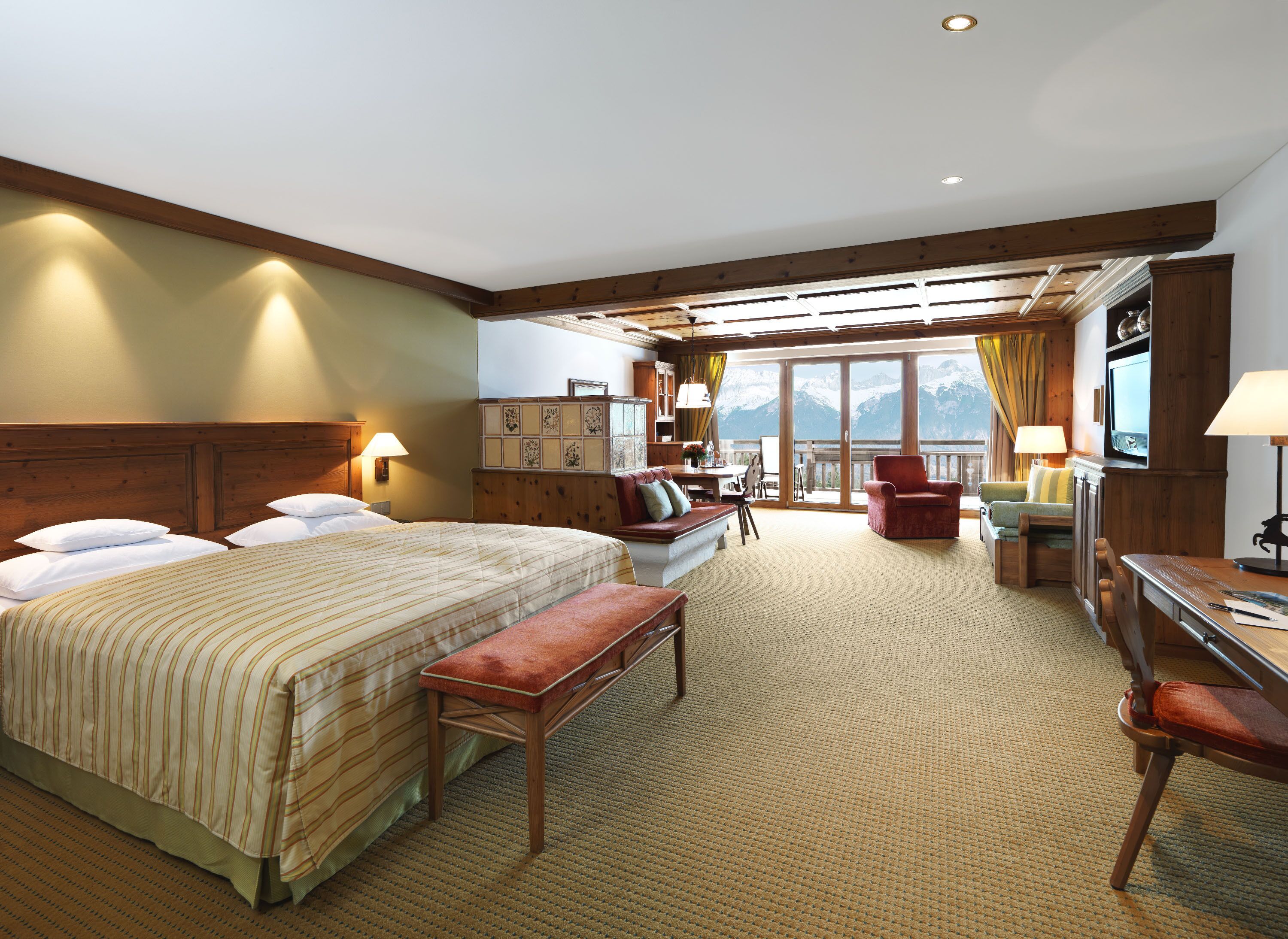 A hotel room with a large bed, a desk, a chair, a sofa, and a balcony with a mountain view.