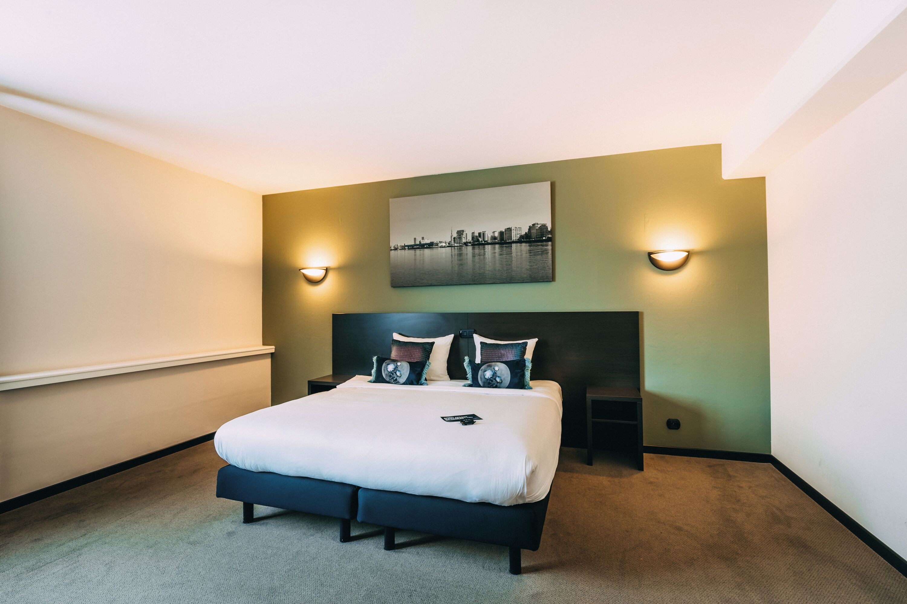 A hotel room with a bed, two bedside tables, a headboard, a wall-mounted picture, and two wall sconces.