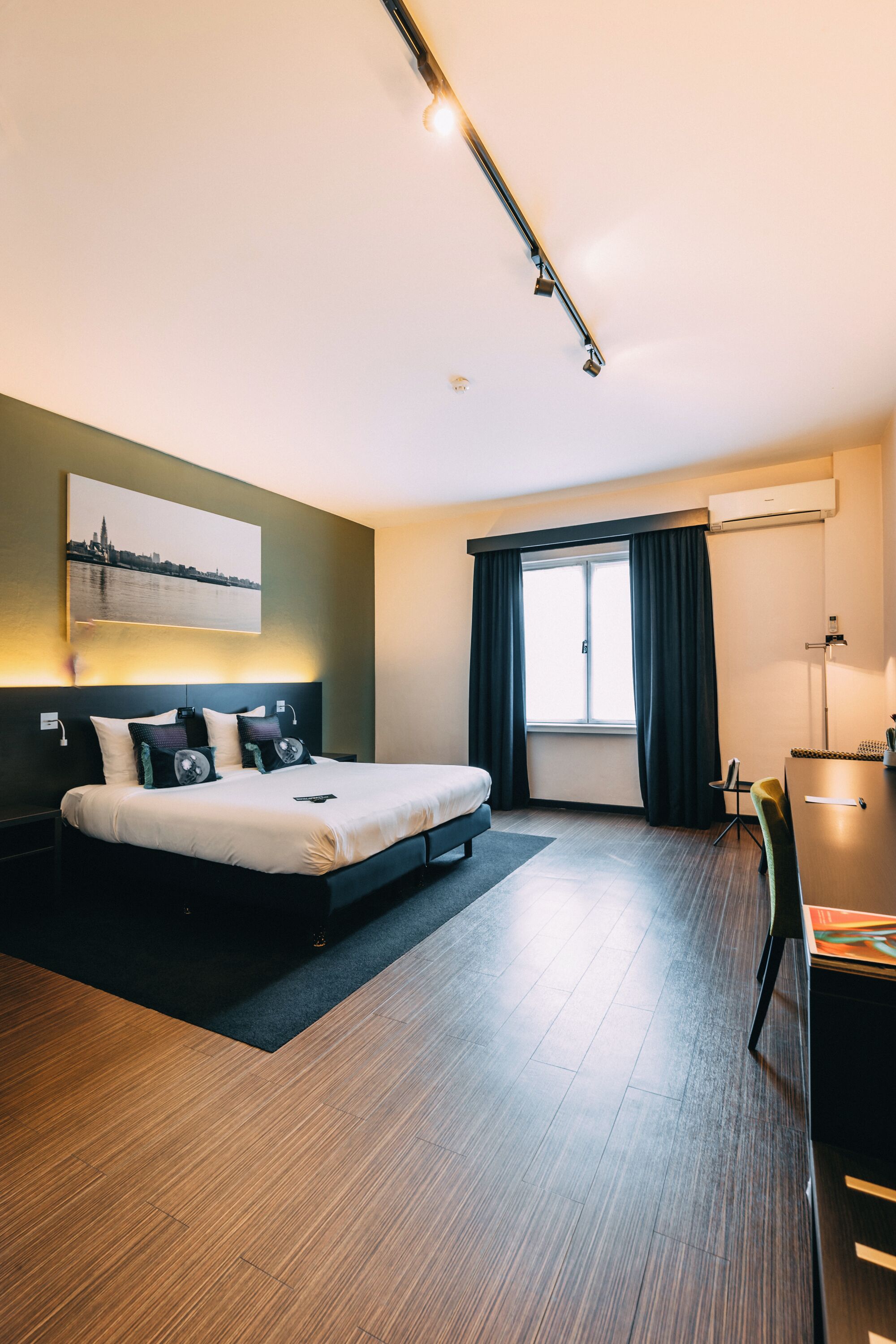Una habitación de hotel moderna con cama, escritorio, silla, ventana con cortinas y un cuadro de un paisaje urbano en la pared.