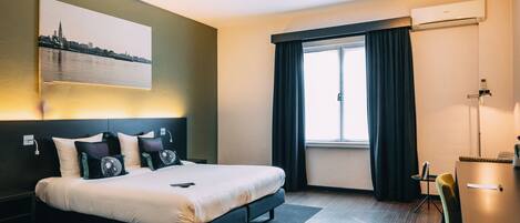 Una habitación de hotel moderna con cama, escritorio, silla, ventana con cortinas y un cuadro de un paisaje urbano en la pared.