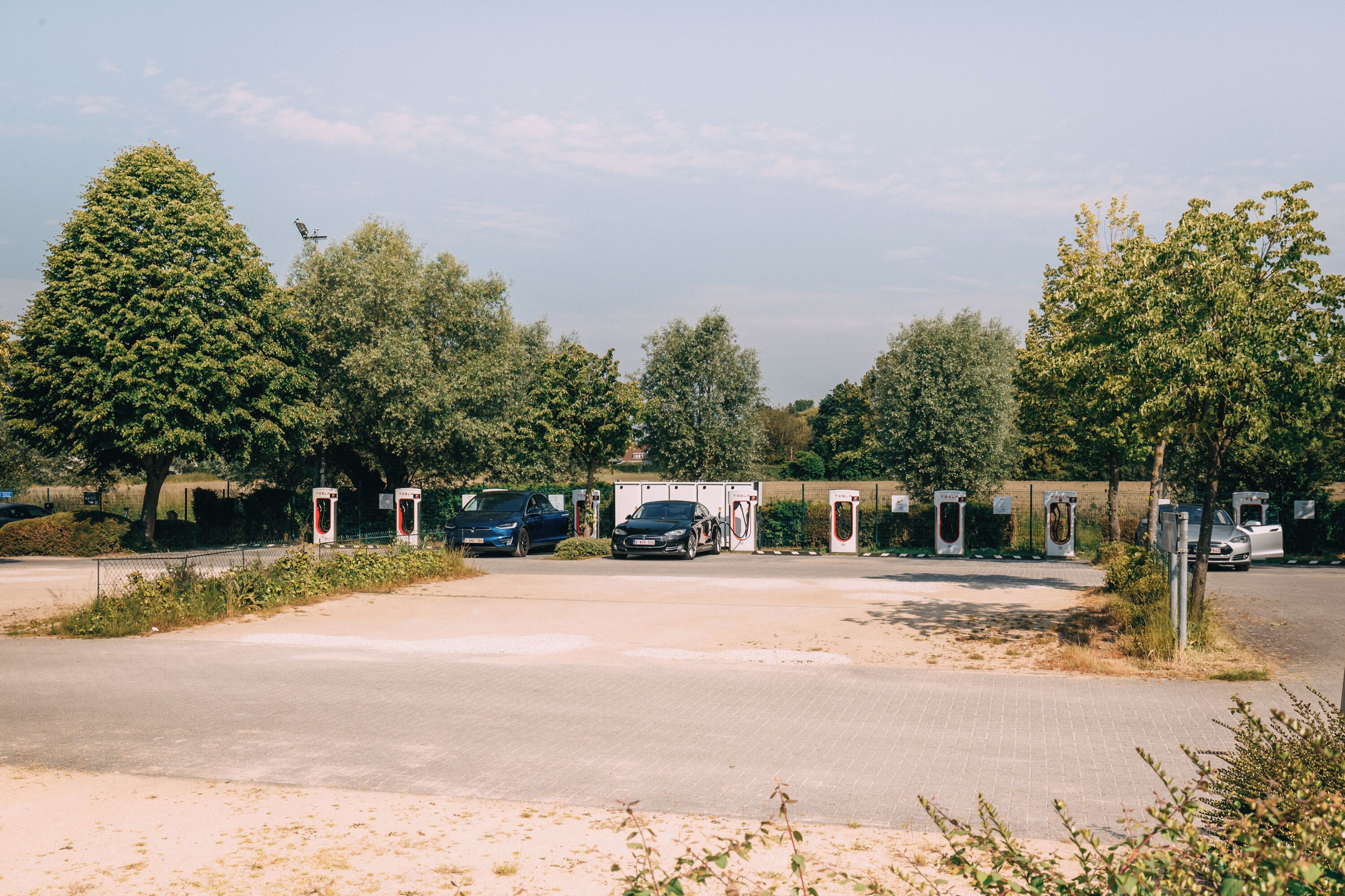 A parking area with electric vehicle charging stations, surrounded by trees and a clear sky.