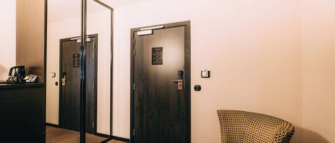 Habitación de hotel con un espejo grande, una puerta de madera oscura, una mesita con cafetera y un sillón con estampado.