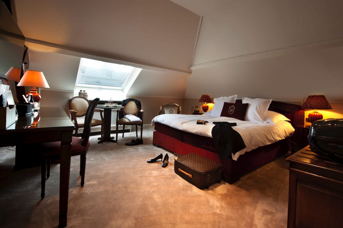 Une chambre d’hôtel avec un lit, un bureau, une chaise et une petite table. Il y a une fenêtre donnant sur le ciel.