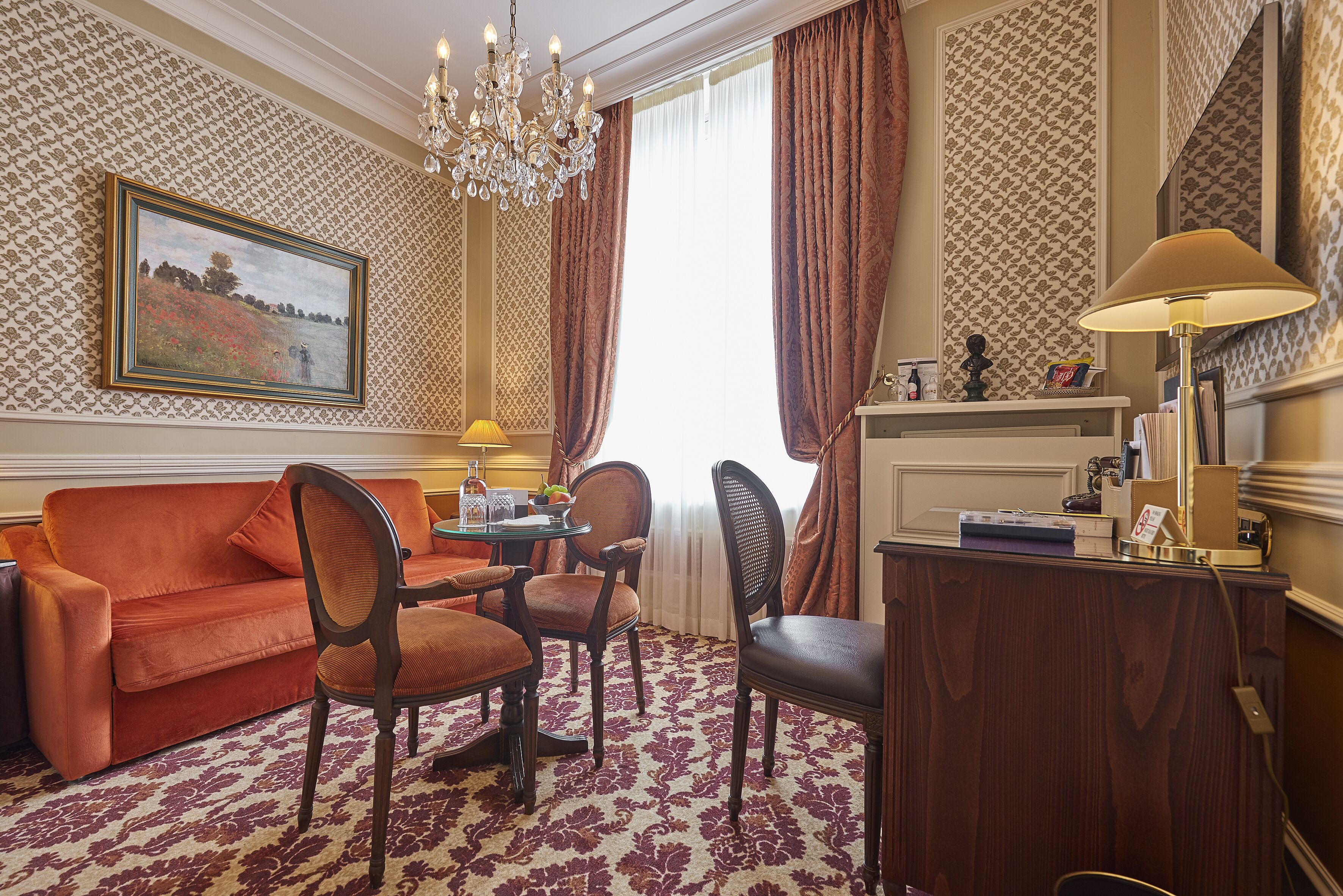 A room with a red sofa, wooden chairs, a small round table, a chandelier, a desk with a lamp, and patterned wallpaper.
