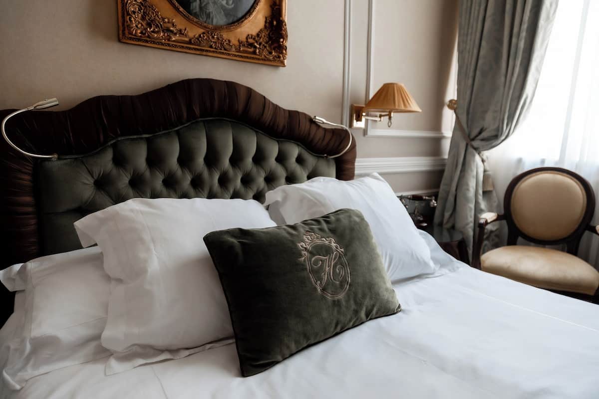 Un lit bien fait, avec une tête de lit sombre, des oreillers blancs et un coussin vert décoratif orné d’un monogramme brodé.