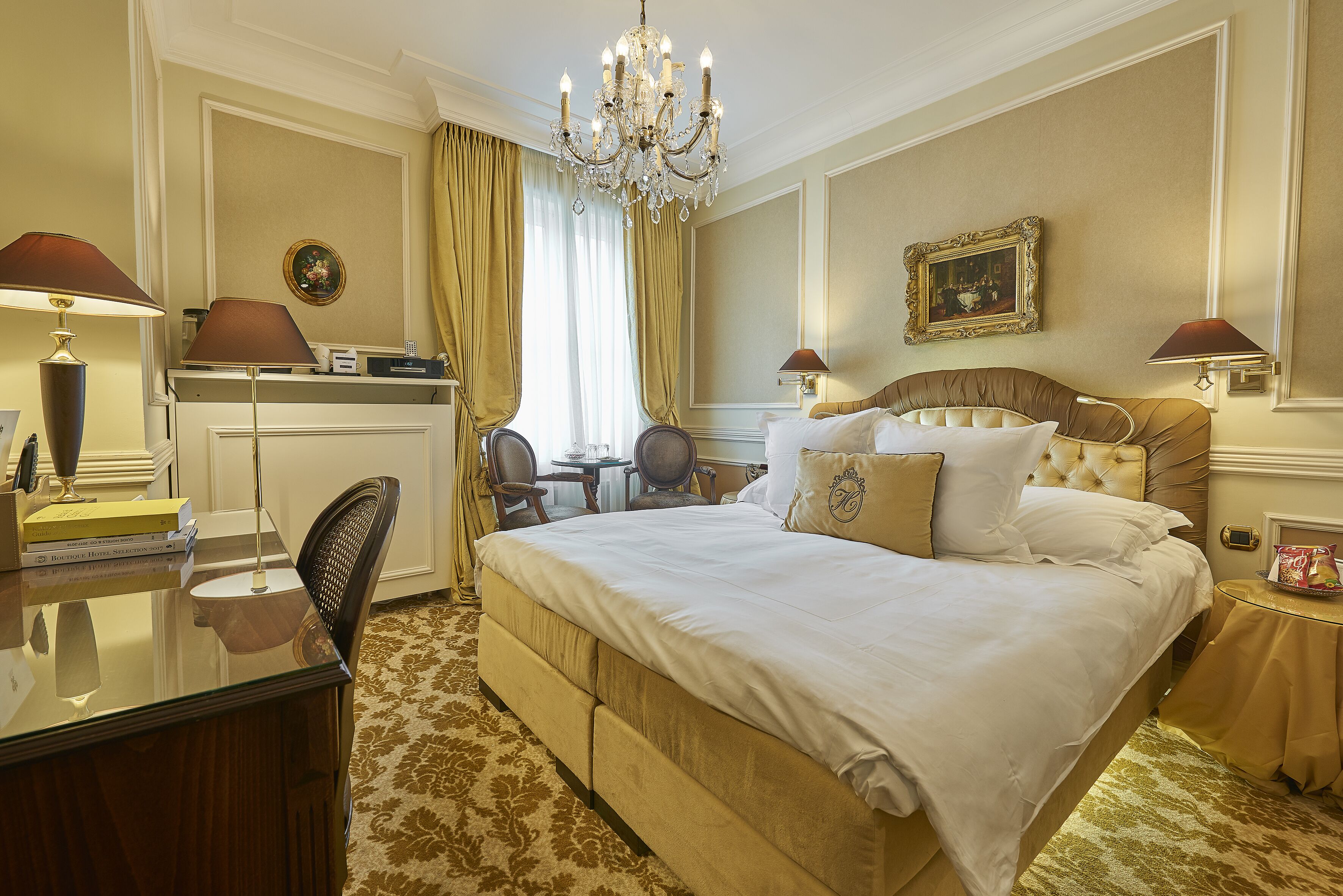 A hotel room with a large bed, a desk, a chair, a chandelier, and a painting on the wall.