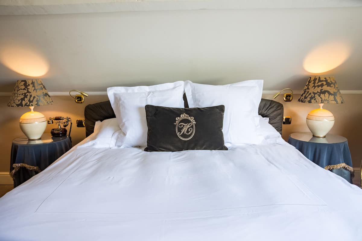 Un lit bien fait, avec du linge de lit blanc, deux oreillers noirs ornés d’un monogramme, et deux tables de chevet avec des lampes.