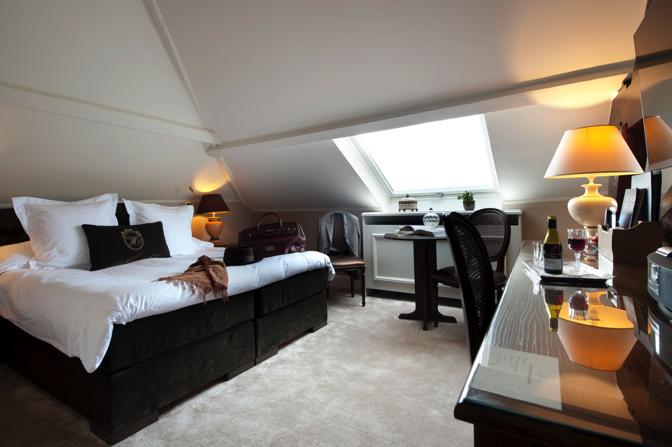 A bedroom with a bed, bedside table, lamp, chair, and a small table with a bottle and glasses.