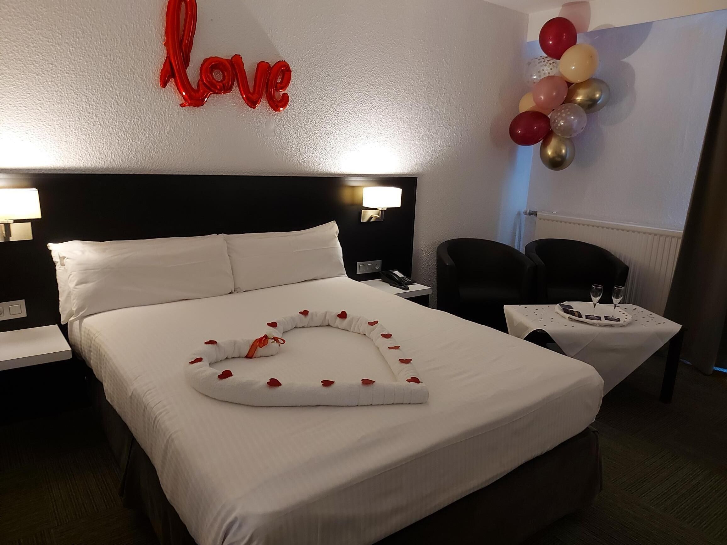 Habitación de hotel con una cama bien hecha, una decoración con forma de corazón sobre la cama, una pared con la palabra 'love' en letras rojas y una mesa con dos vasos y un menú.