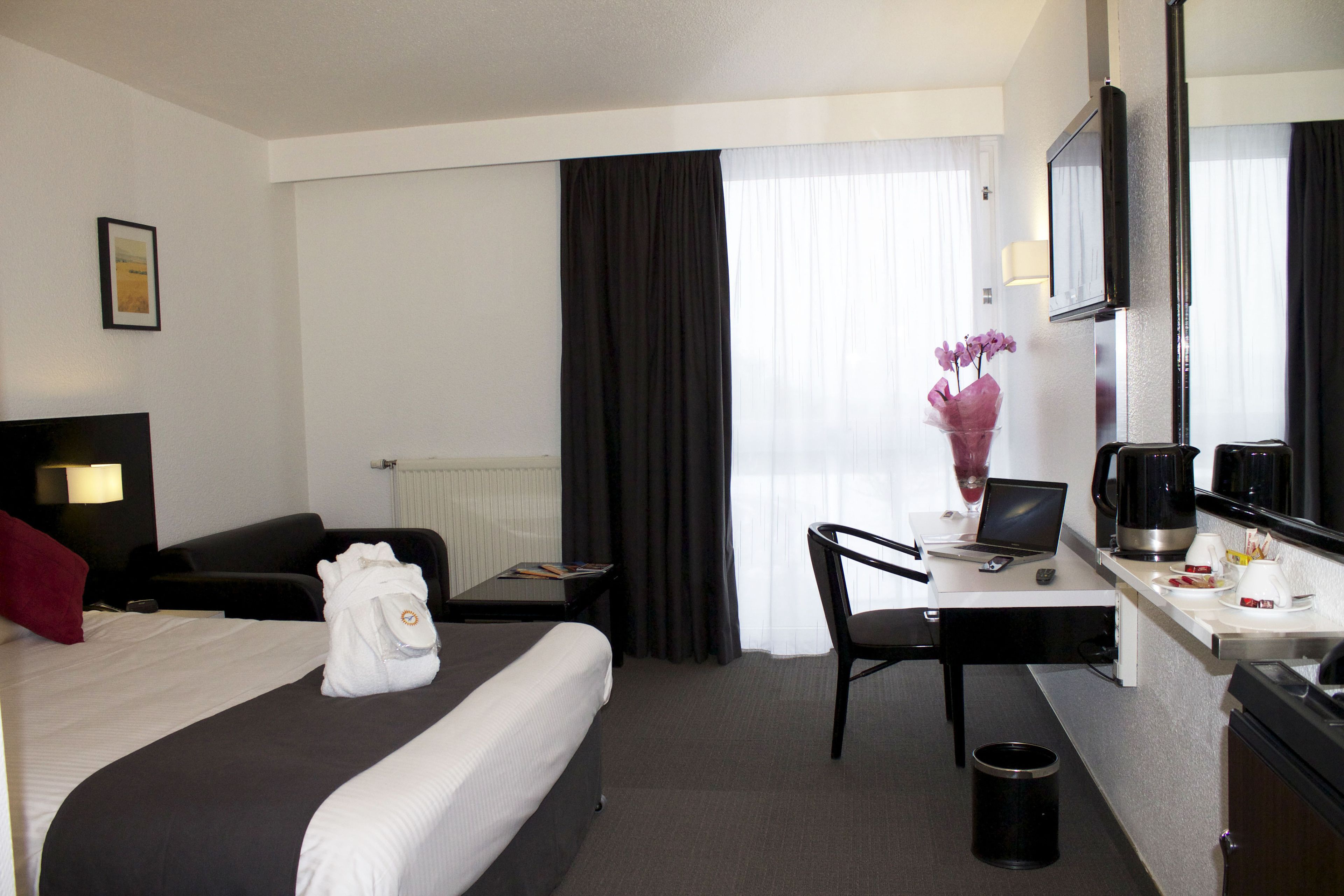 A hotel room with a bed, a desk with a laptop, a chair, a coffee maker, and a trash can.