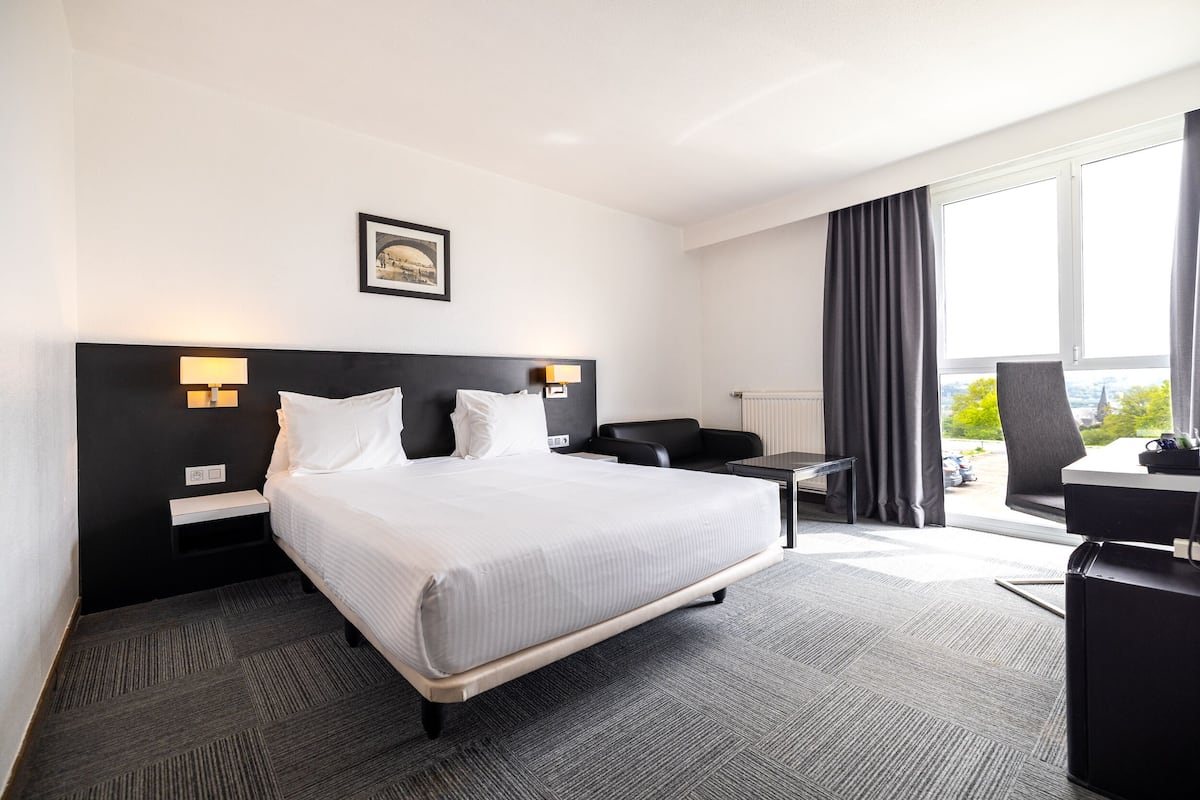 Ein Hotelzimmer mit Bett, Nachttischen, Schreibtisch, Stuhl und einem fensterbepflanzten Fenster.
