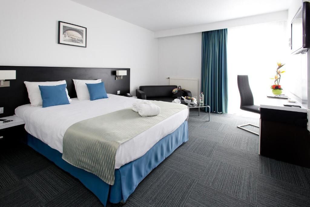A hotel room with a large bed, a desk, a chair, a TV, and a window with curtains.