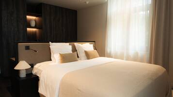 A hotel room with a large bed, a bedside table with a lamp, a wall-mounted shelf with a vase, and a window with sheer curtains.