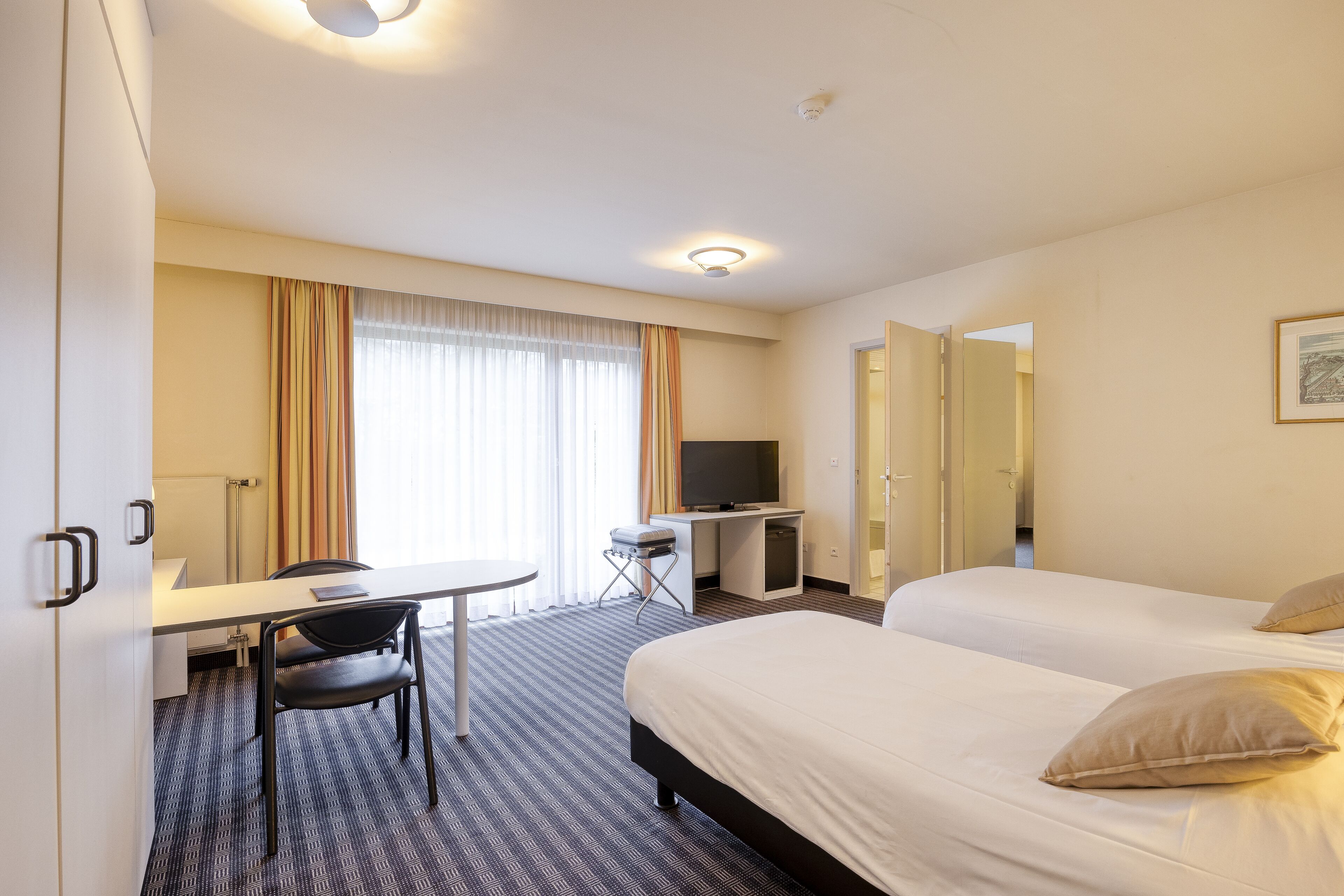 Habitación de hotel con cama, escritorio, silla, televisor y ventana con cortinas.
