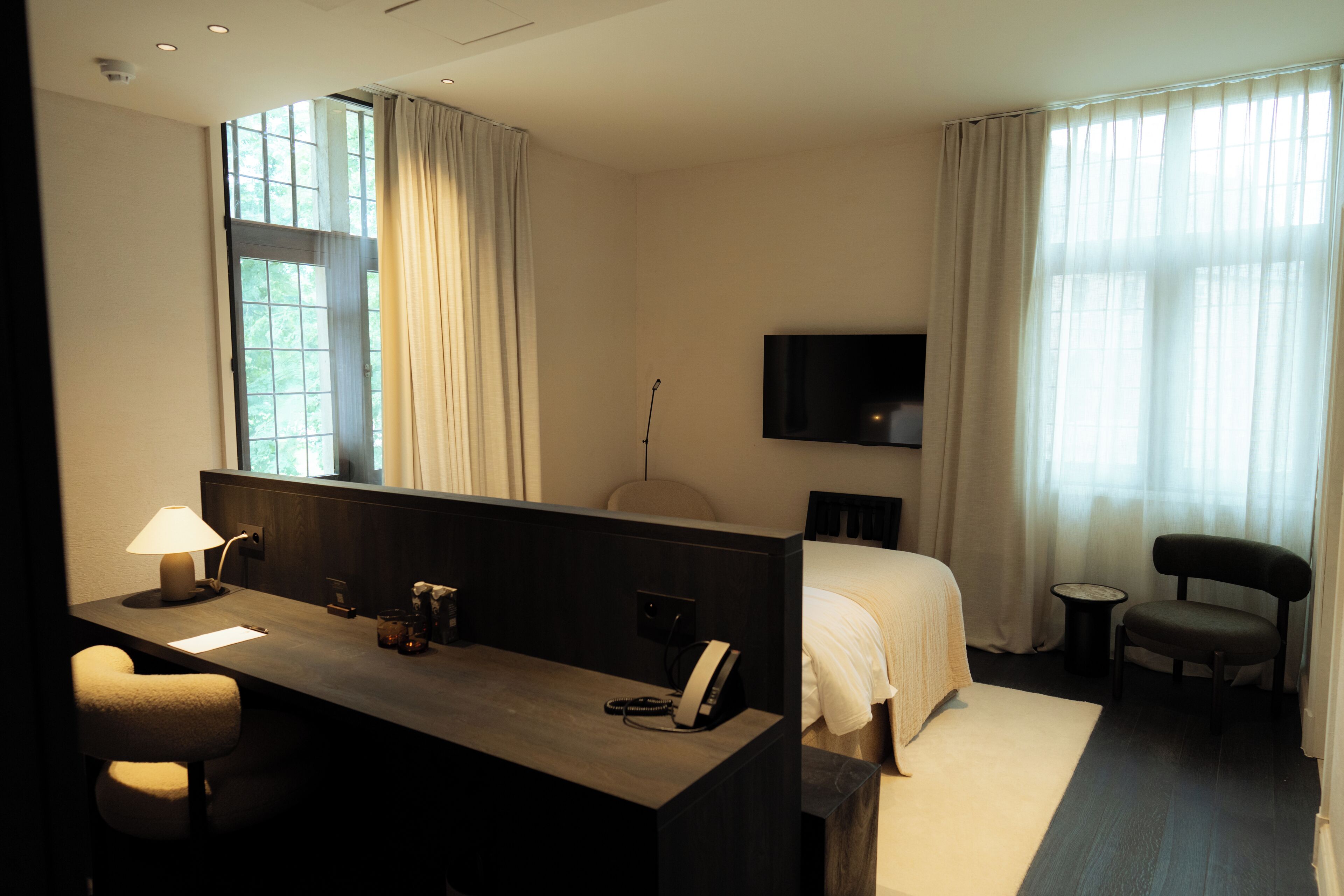 A hotel room with a desk, chair, bed, TV, and large windows with curtains.