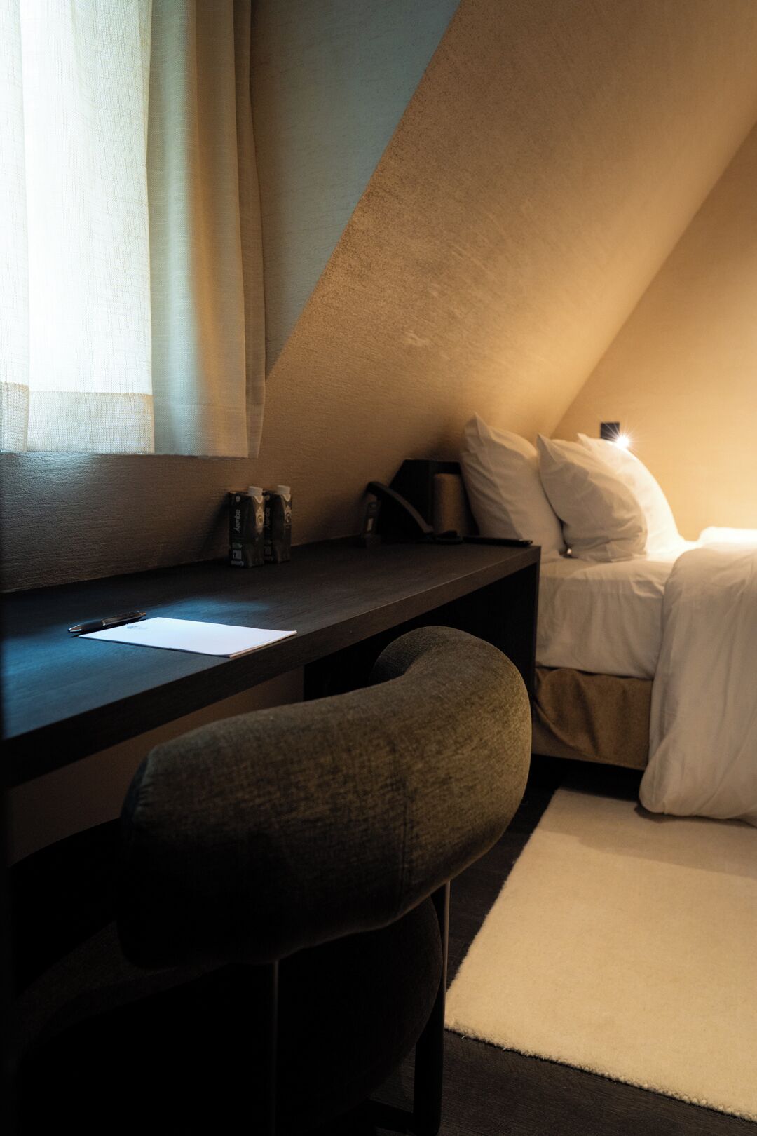 A small, dimly lit room with a desk, a chair, a bed, and a window with curtains.