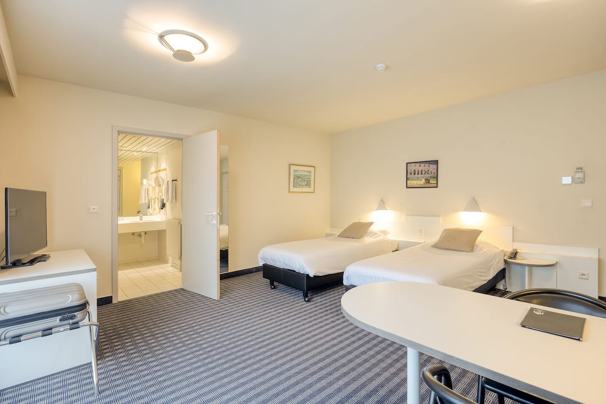 A hotel room with two beds, a desk, a chair, a TV, and a bathroom visible through an open door.
