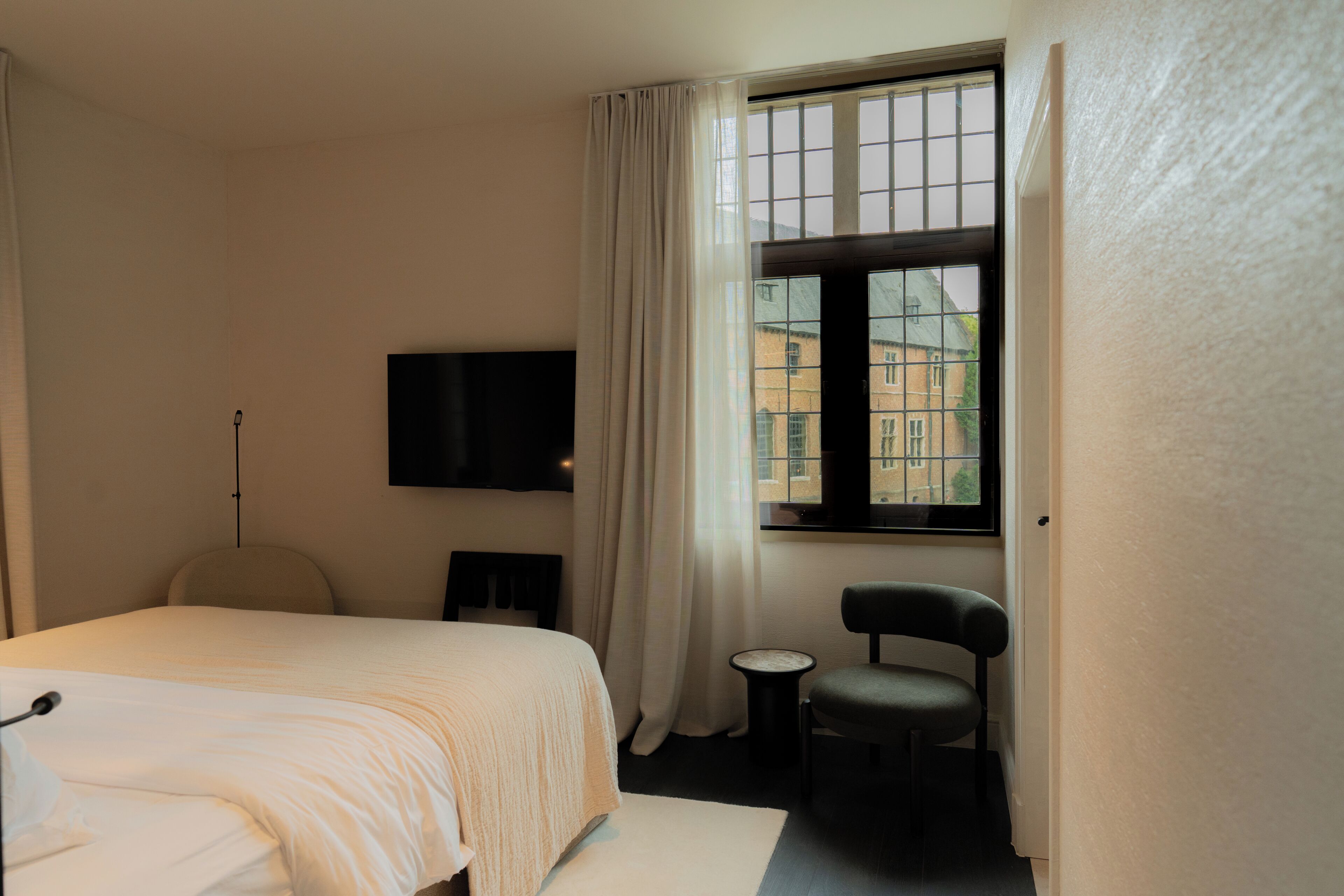 A hotel room with a bed, a chair, a small table, a television, and a window with curtains.