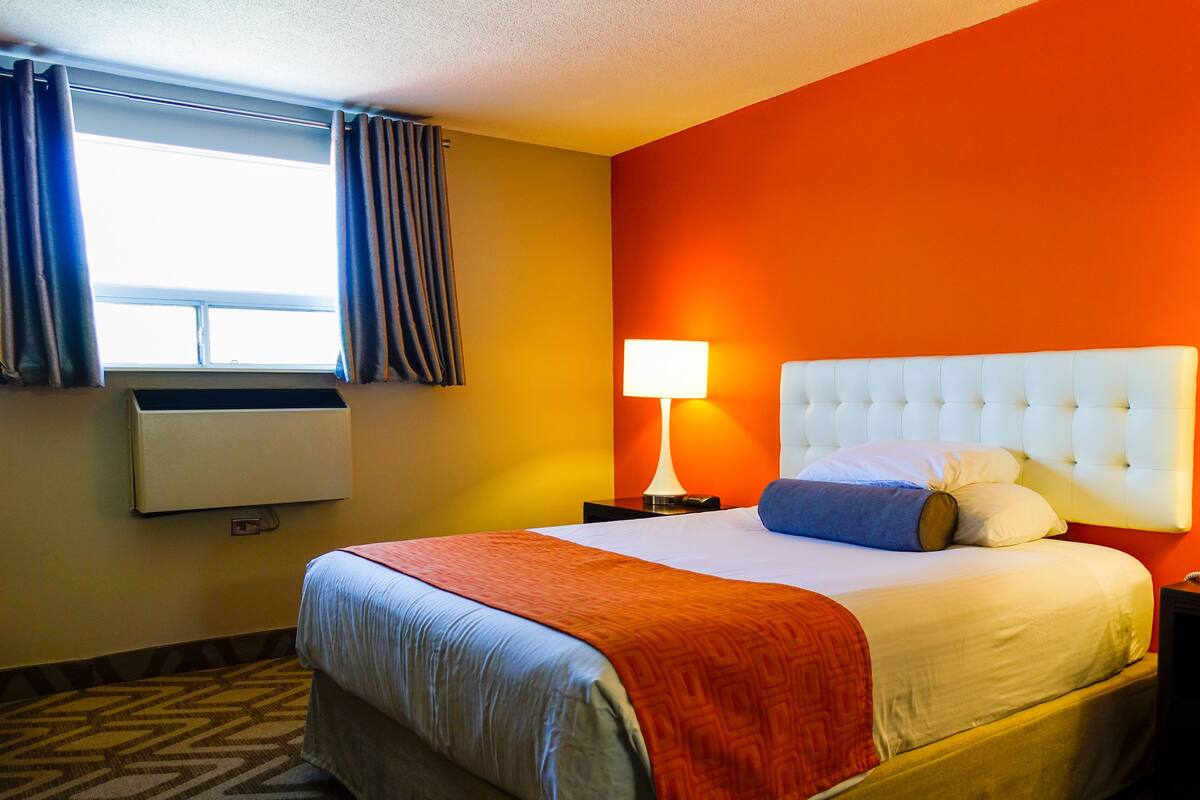 Una habitación de hotel con cama, mesita de noche, lámpara, aire acondicionado y ventana con cortinas.
