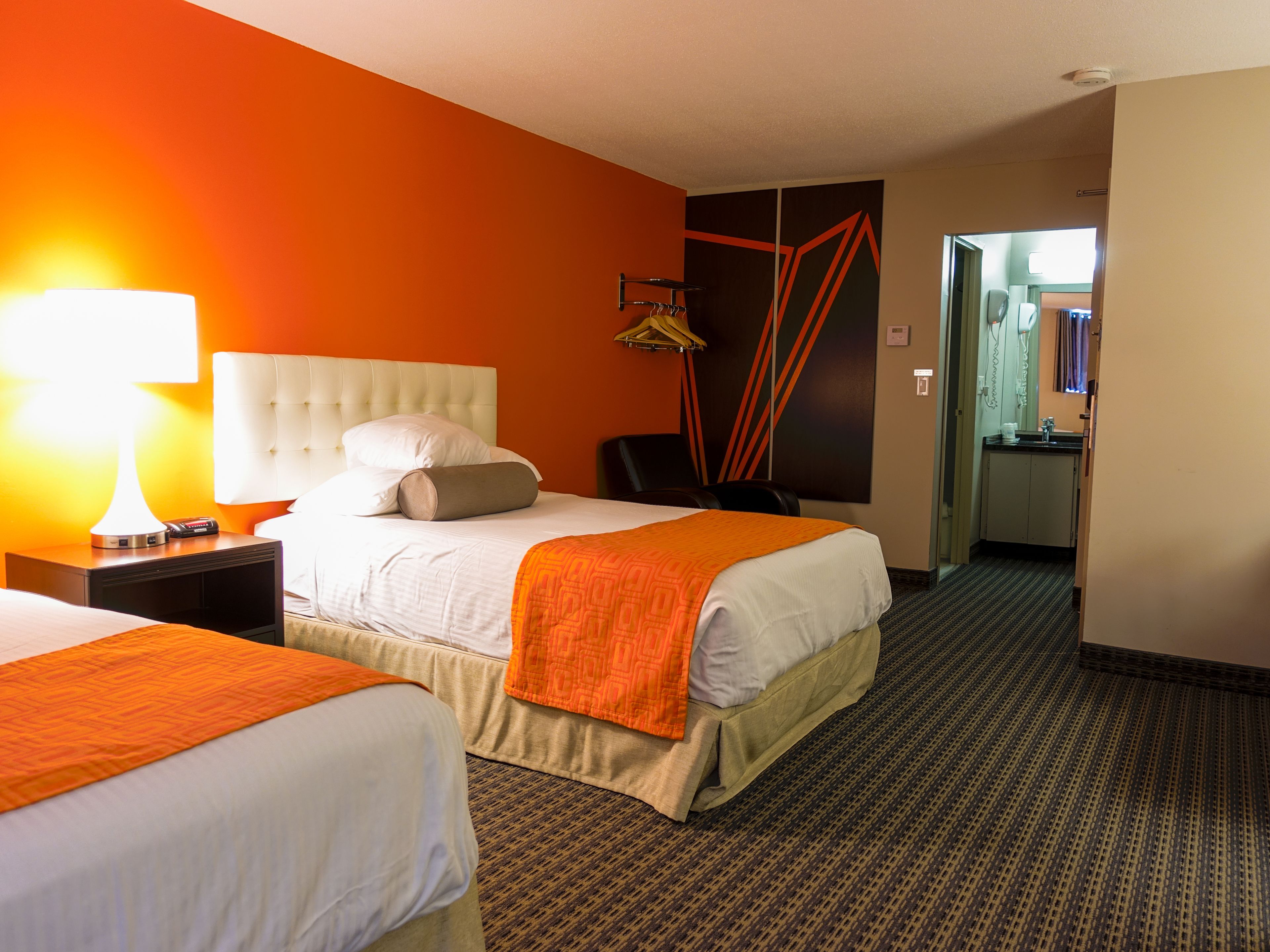 A hotel room with two beds, a nightstand with a lamp, a wardrobe, and a bathroom visible through an open door.
