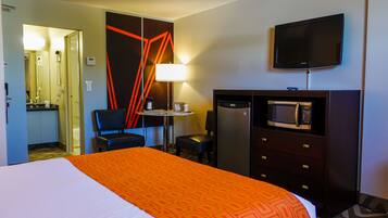 A hotel room with a bed, a desk with a lamp, a microwave, a refrigerator, and a television.